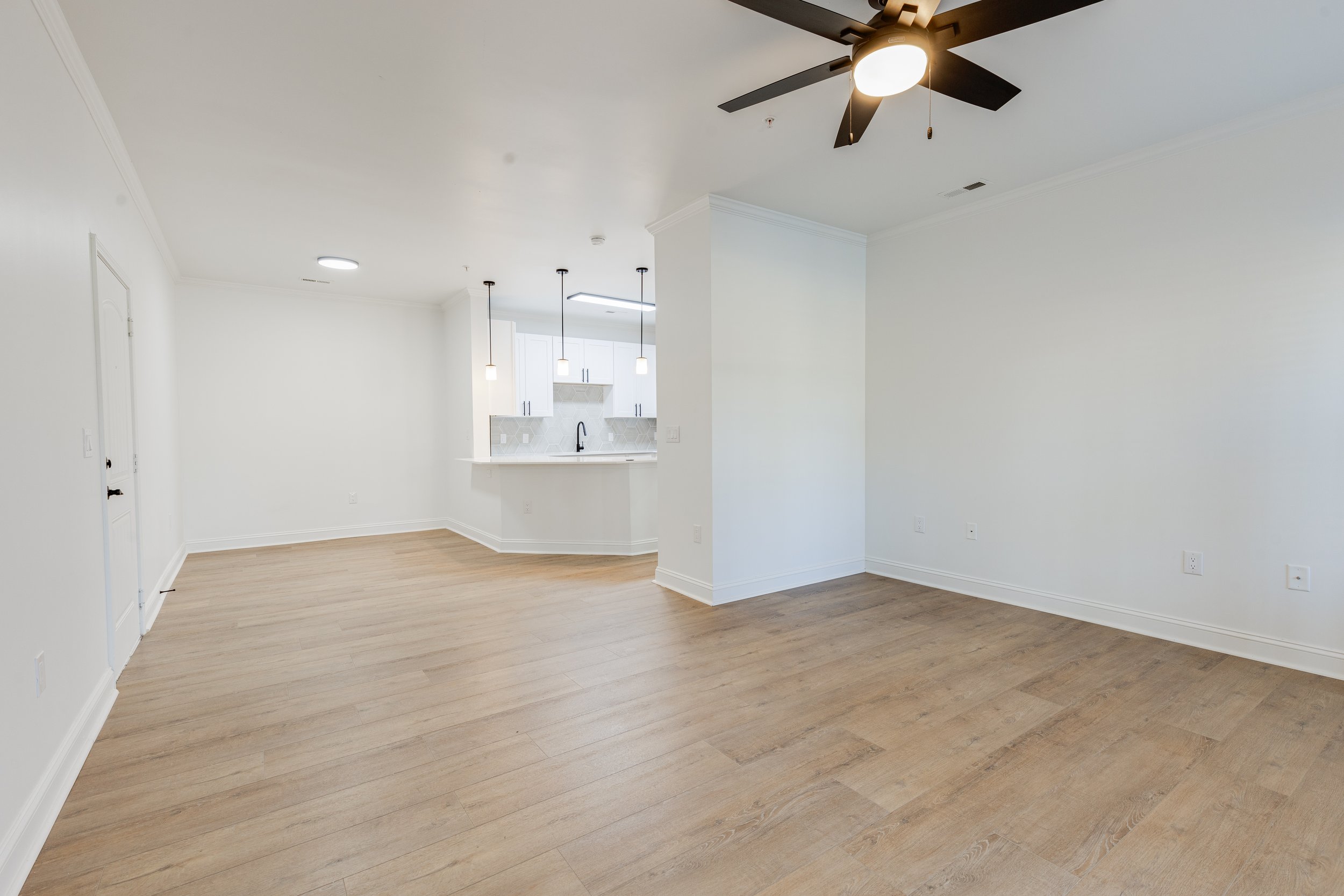 Empty living room with light wood flooring, white walls, and a ceiling fan, adjacent to a kitchen with white cabinets and hanging pendant lights.