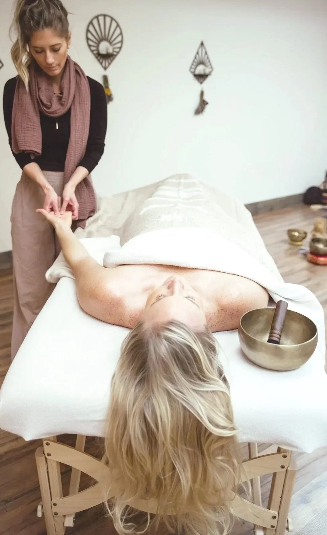 A woman lying face up on a massage table, receiving a massage from a therapist. The scene is held in Spirit Alchemy Studio with decorative wall hangings.