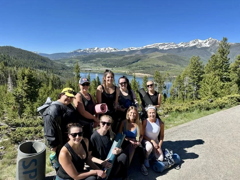 Alison providing a private yoga session for a group of women outdoors with a scenic background of a lake, forest, and snow-capped mountains on a clear, sunny day.