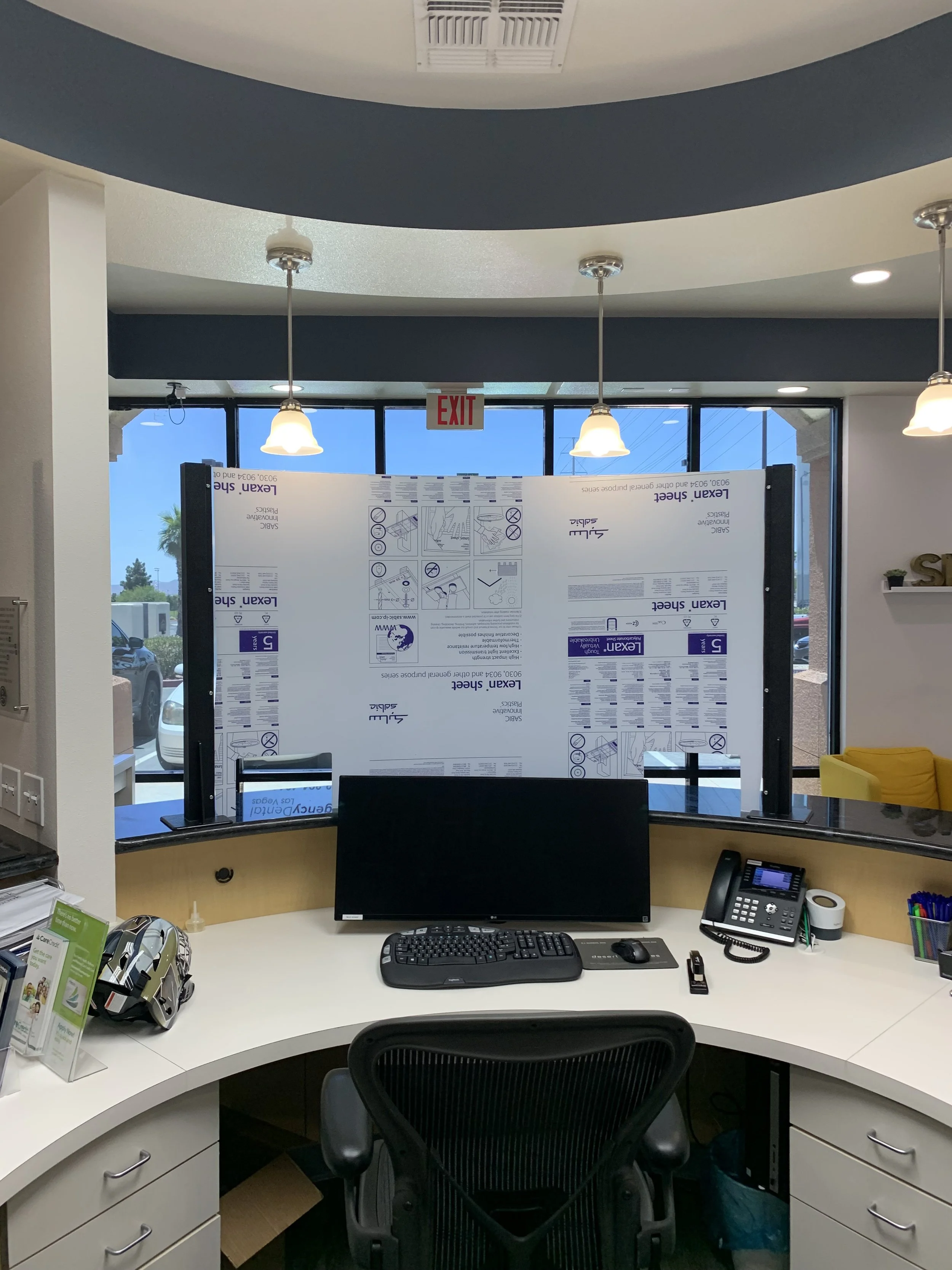 A reception desk with a computer monitor, keyboard, mouse, desk phone, and office supplies. Behind the desk is a partition with instructions and notices. There is a window with a view of the outdoors, and ceiling lights hang above the desk.