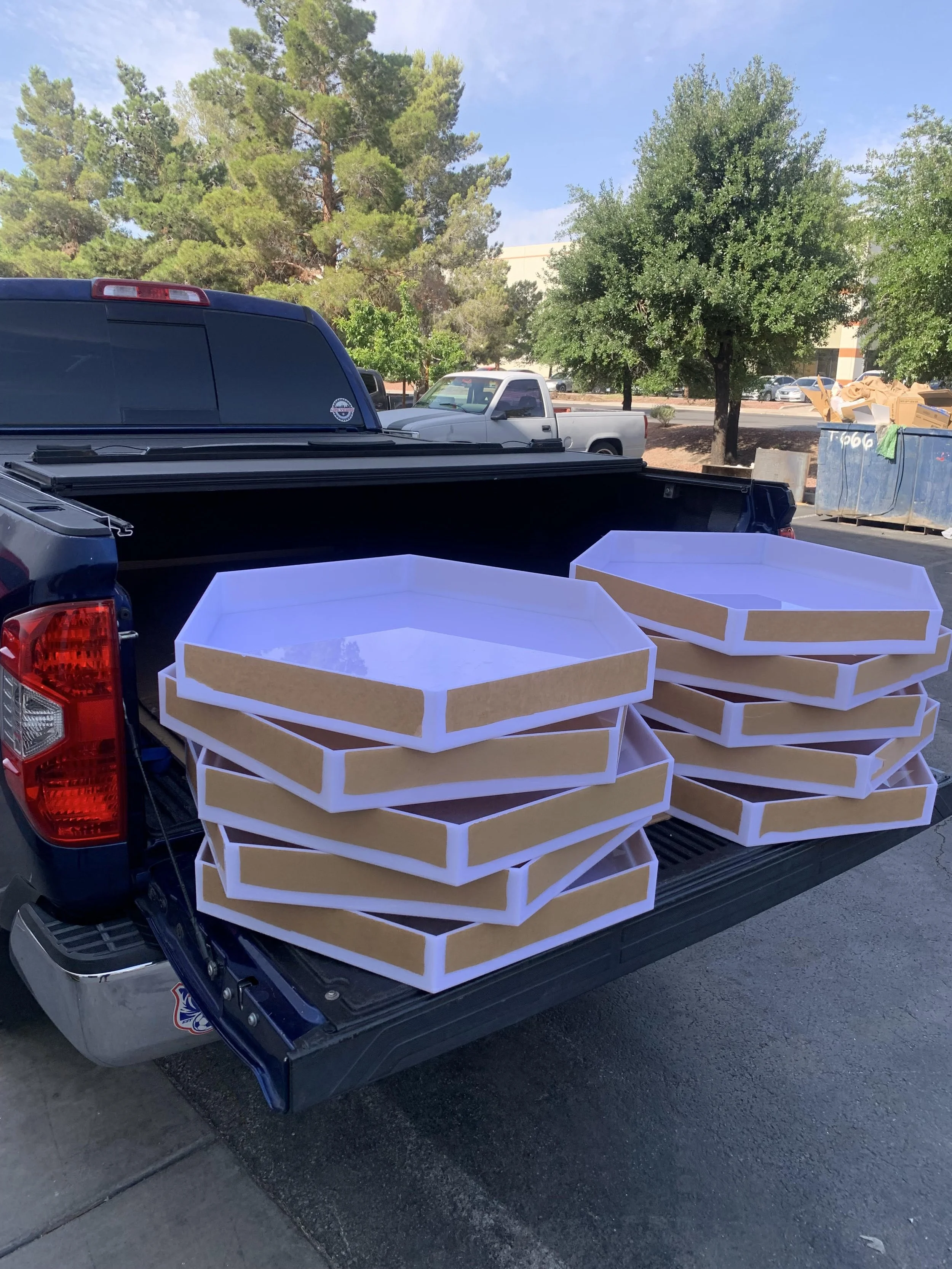 Pickup truck with empty low-sided boxes in the back, parked in a parking lot near trees and a dumpster.