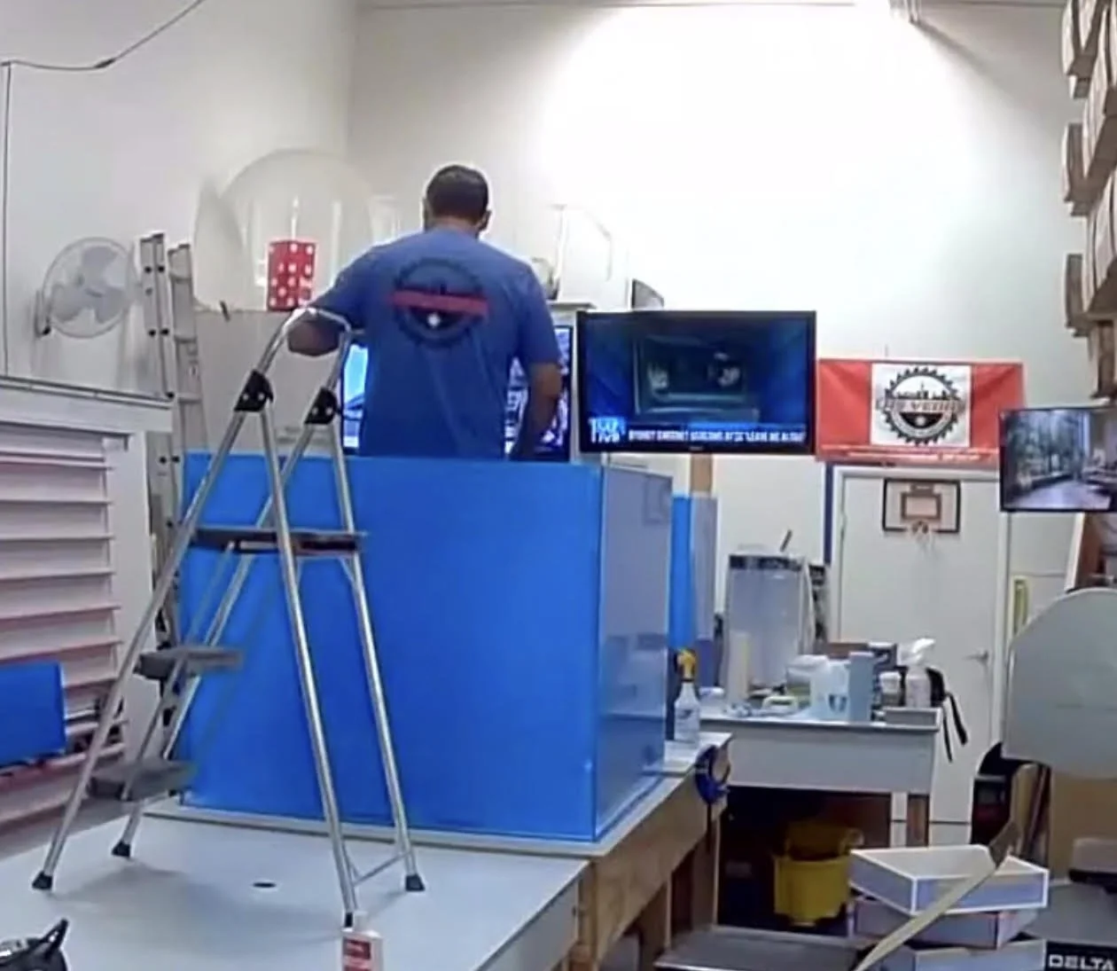 A man with a prosthetic arm working in a workshop with shelves, a ladder, and various tools and supplies around him. There is a TV and a flag on the wall.