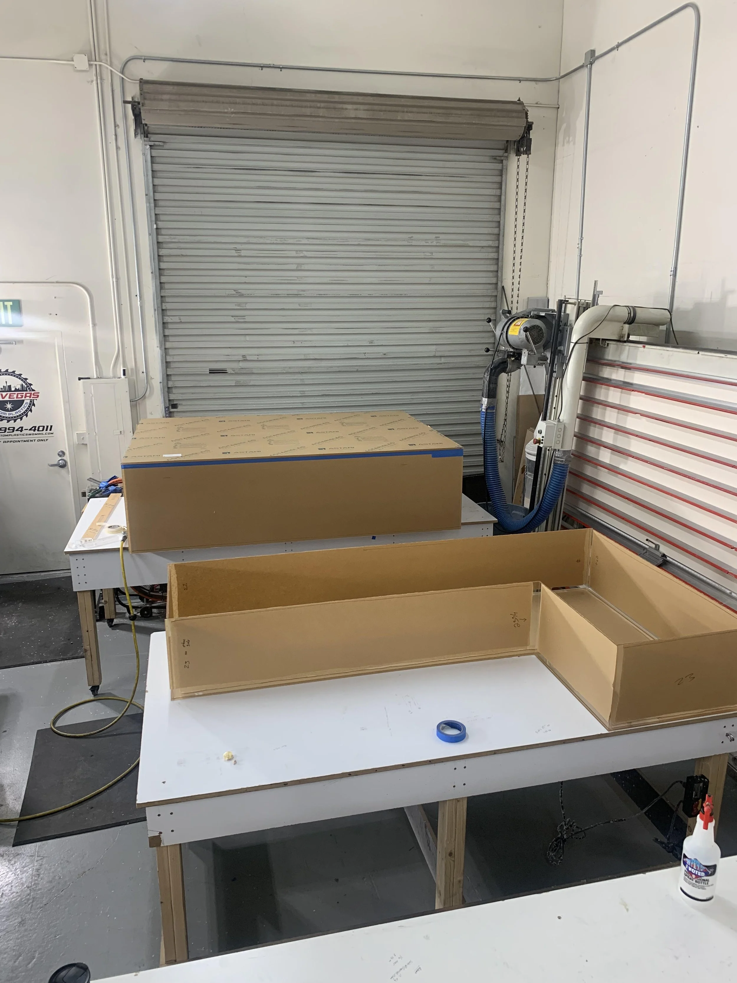 Workshop with tables, cardboard boxes, tools, and a partially open metal roll-up door.