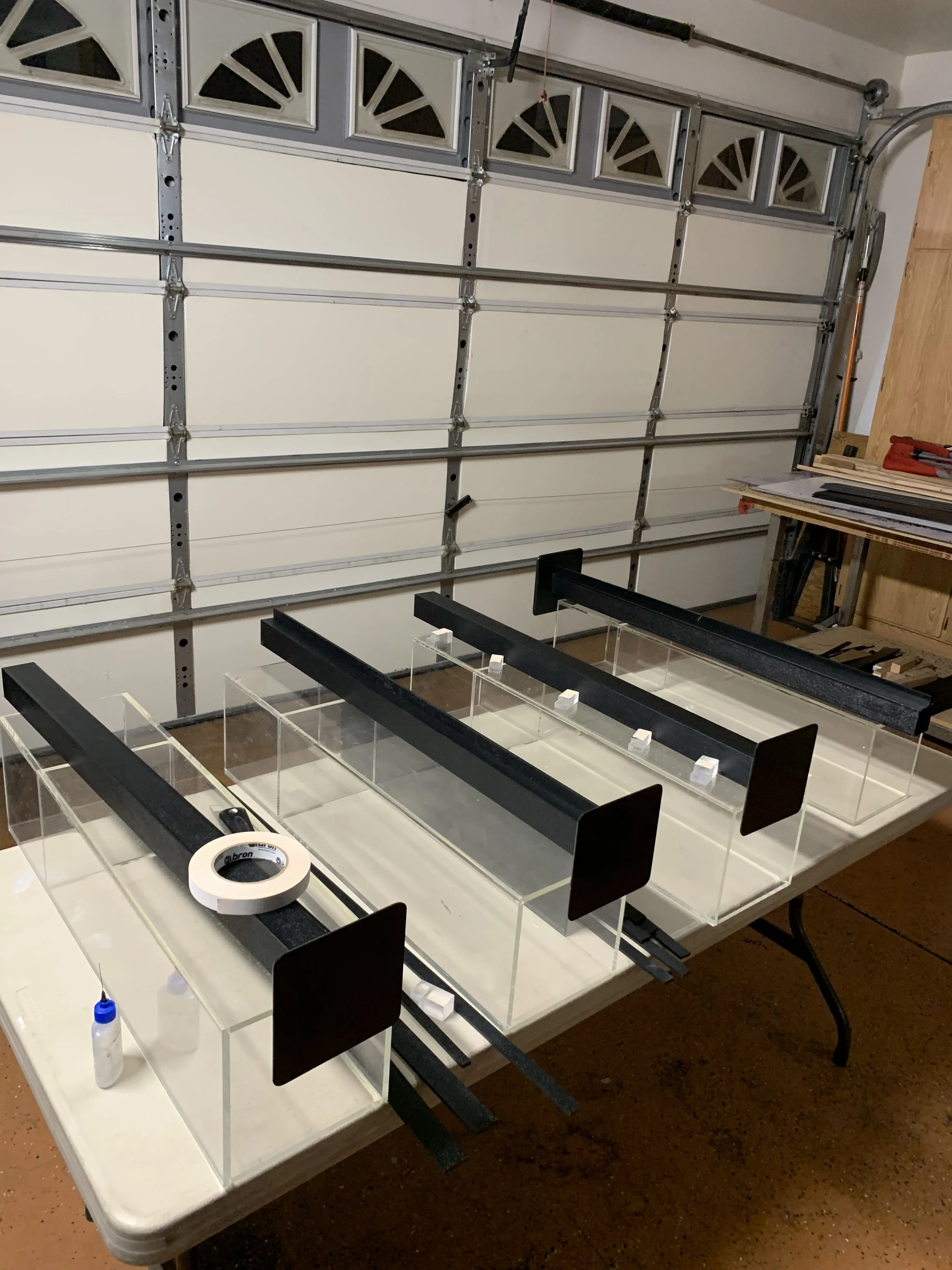 A partially assembled flower display case with a clear glass top and black trim, sitting on a table surrounded by tools and small parts, in a workshop with a garage door in the background.