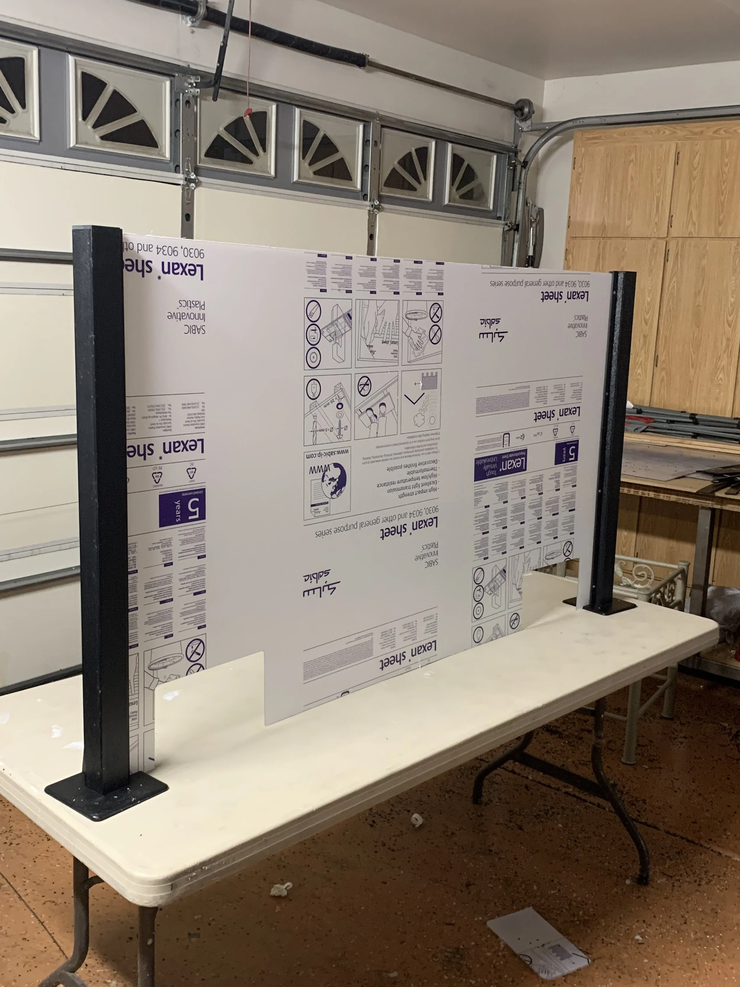 A partially assembled light gray garage door with decorative window panels. In front of it, a work table holds a large boxed item labeled 'Lekan Sheet,' with instructions and diagrams, supported by black metal legs. The floor is brown speckled tile, with a small piece of paper or packaging on it.