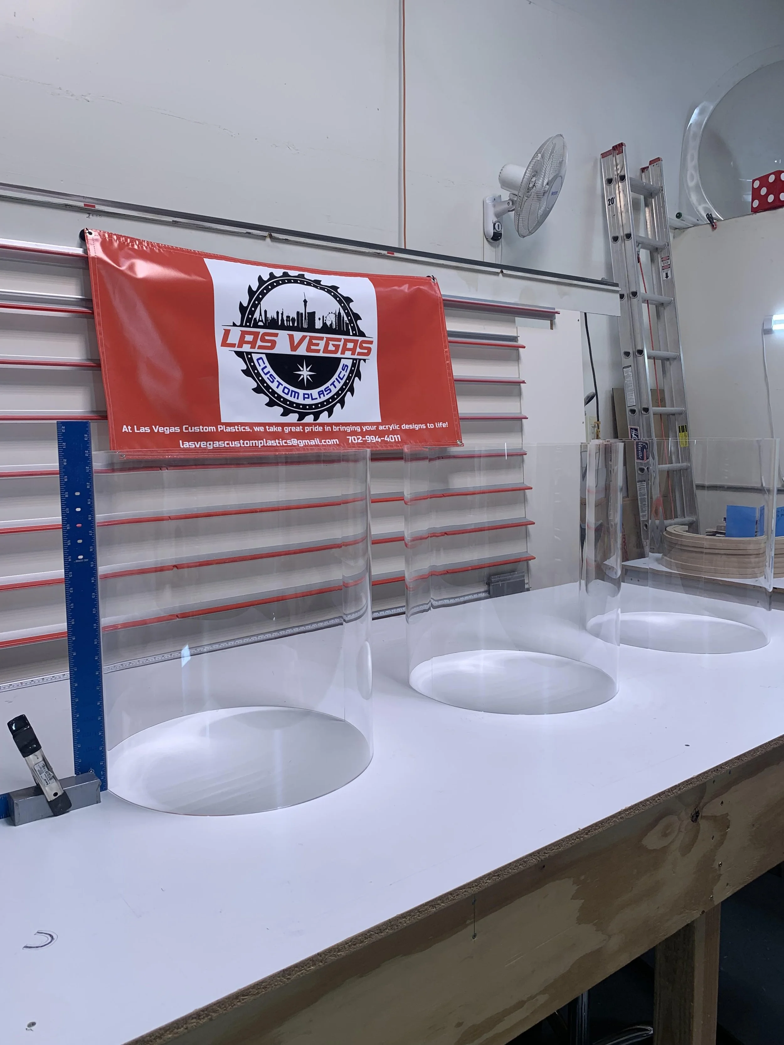Laboratory workbench with three clear plastic cylinders, a blue ruler, and a black marker, with a Las Vegas Custom Plastics banner hanging on the wall behind.