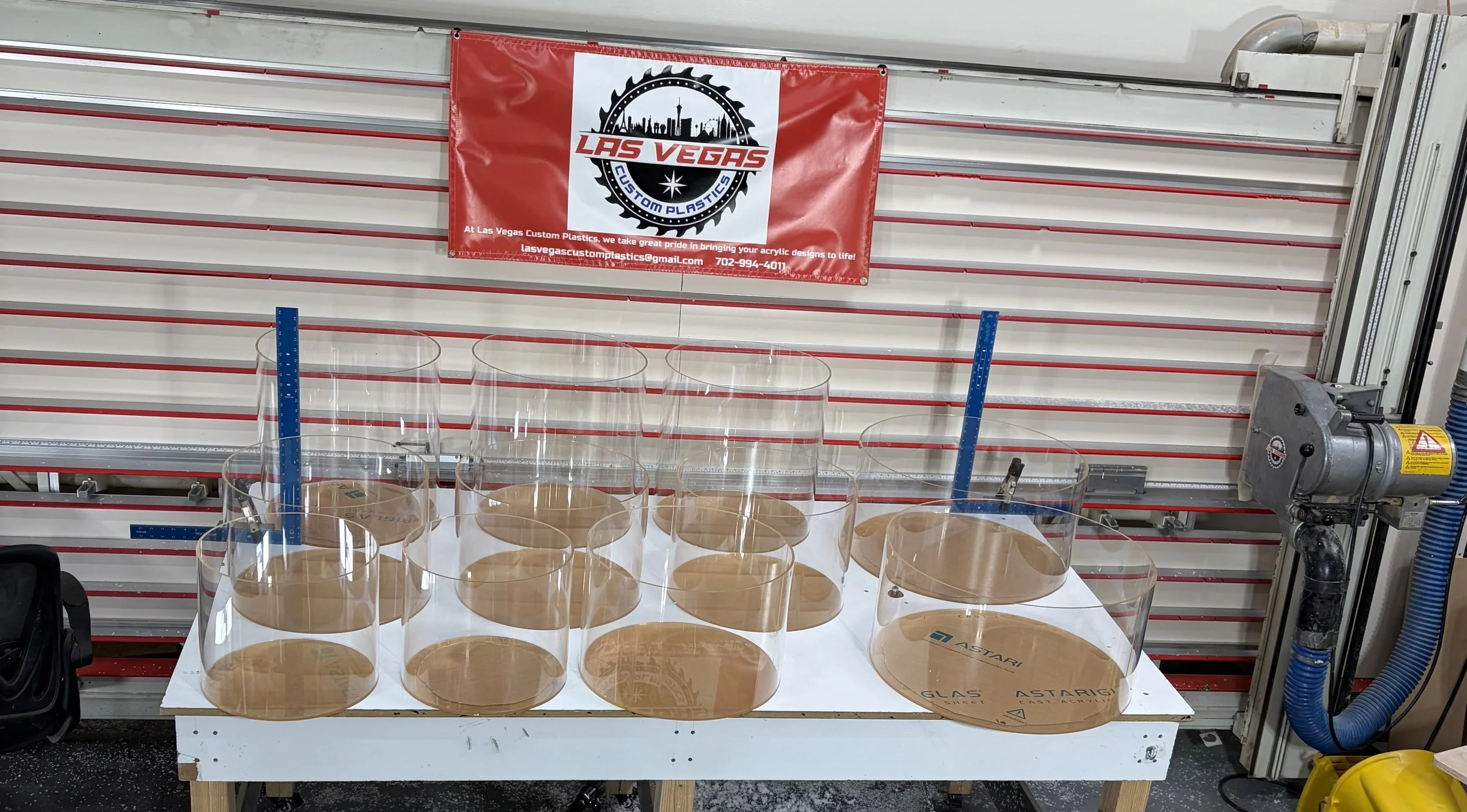 Clear acrylic sheets with circular cutouts arranged on a white table, against a red and white slatted wall with a Las Vegas Custom Plastics banner.