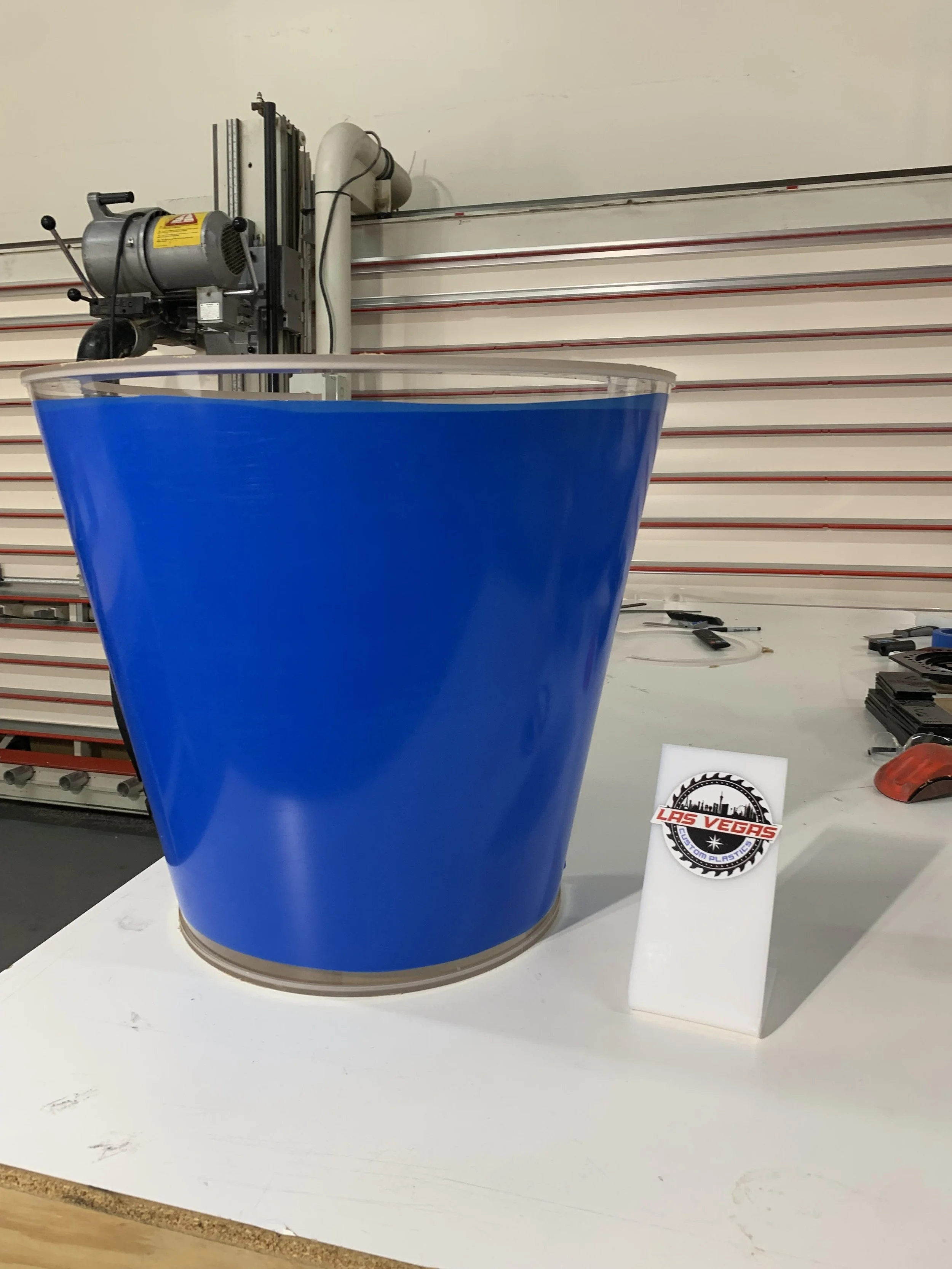 A blue plastic cup with a beige-colored base placed on a white display stand. There are tools and a sticker that reads "Las Vegas custom plastics" on the stand. In the background, there are metal rods and equipment in a workshop setting.