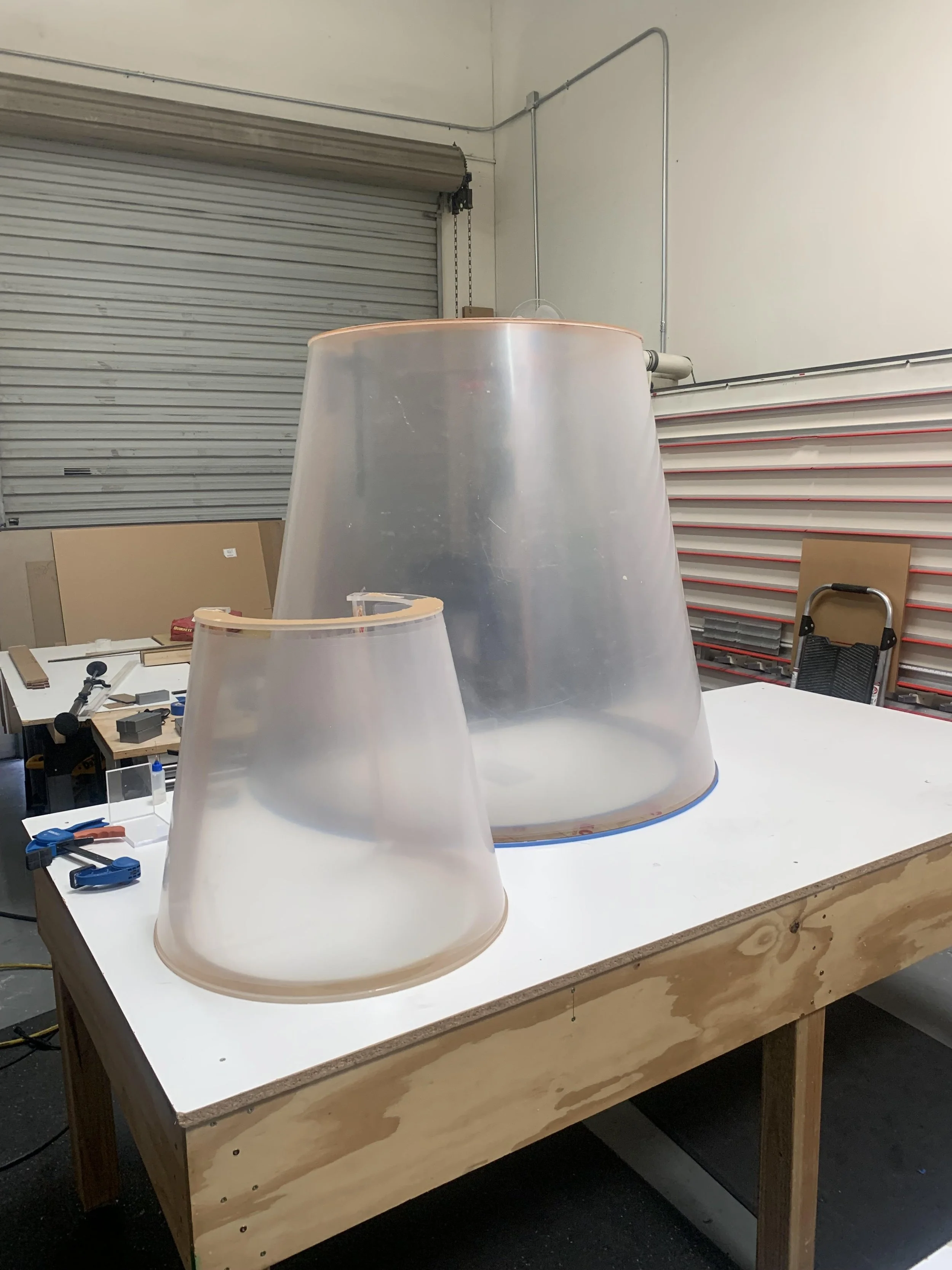 Workshop with two large translucent lampshades on a white table, tools and materials scattered, wooden table frame, background includes a metal roller door, shelving with red rods, and various workshop items.