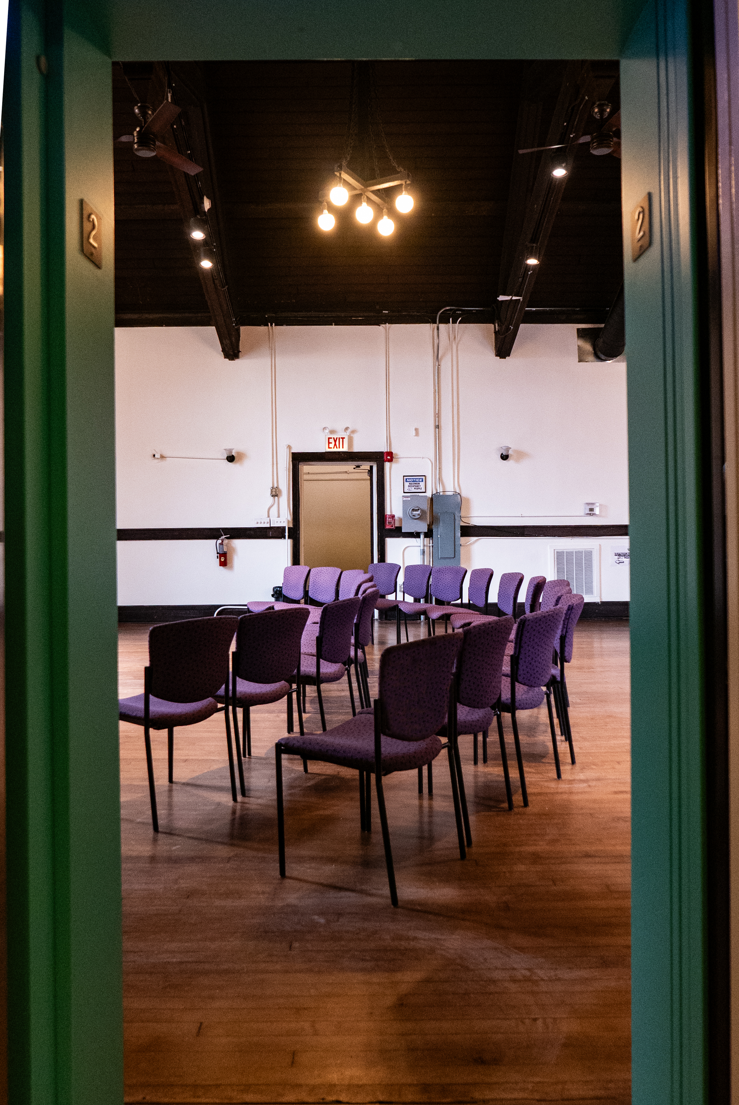 Empty room with purple chairs arranged in rows, white walls, and a dark wooden ceiling with exposed beams and a light fixture, viewed through an open door with green trim.