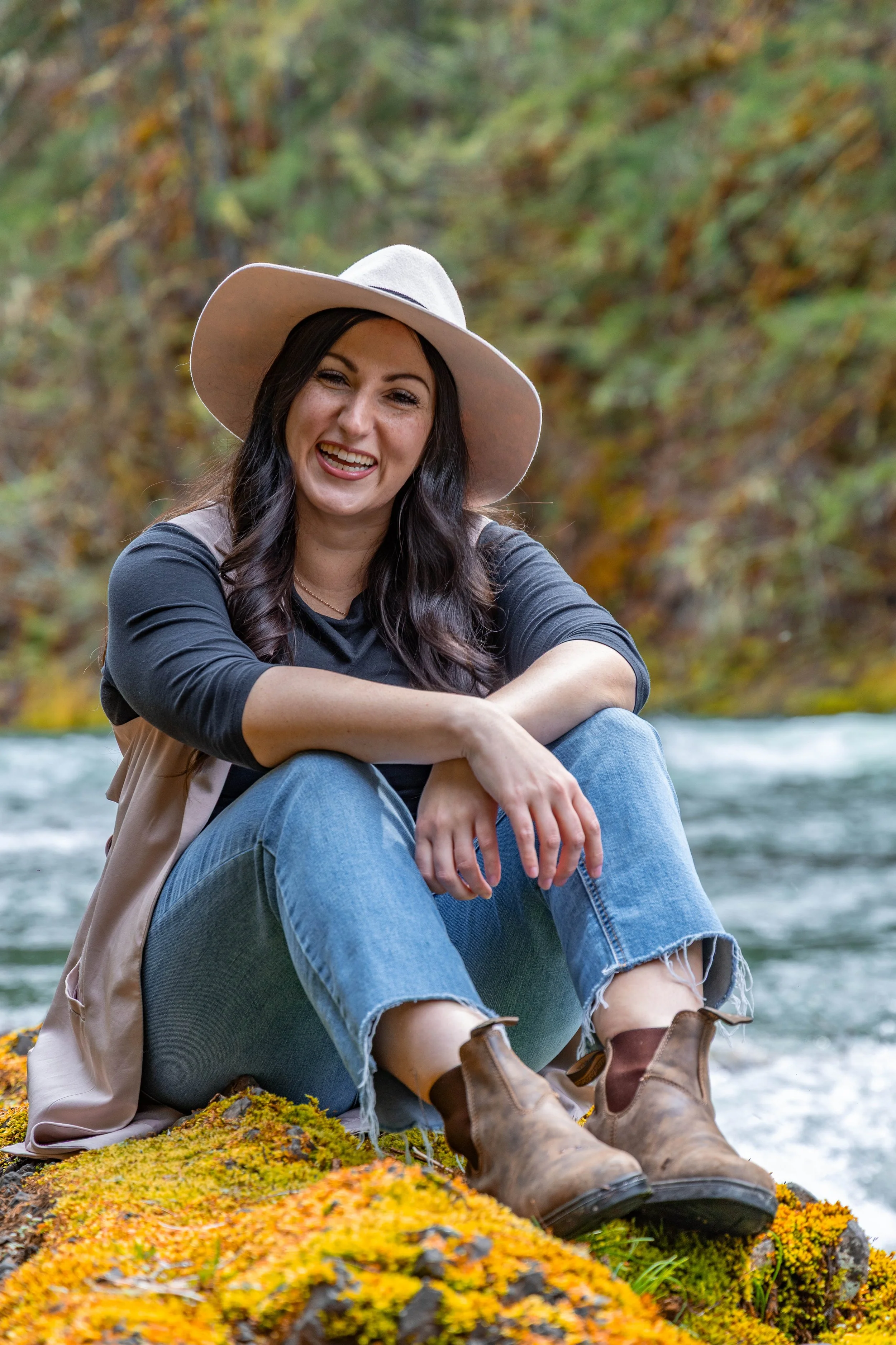 Natural outdoor portrait of A woman sitting on a mossy rock by a river, smiling, wearing a wide brim hat, black shirt, jeans, and brown boots, surrounded by fall foliage Cowichan River BC