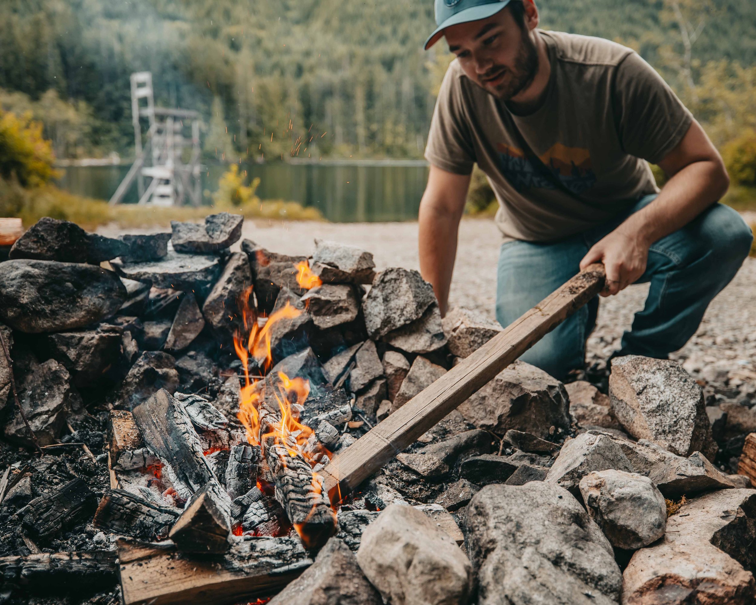 a man in a green shirt and jeans pokes at a blazing campfire with a stick next to a remote backcountry lake on vancouver island british columbia