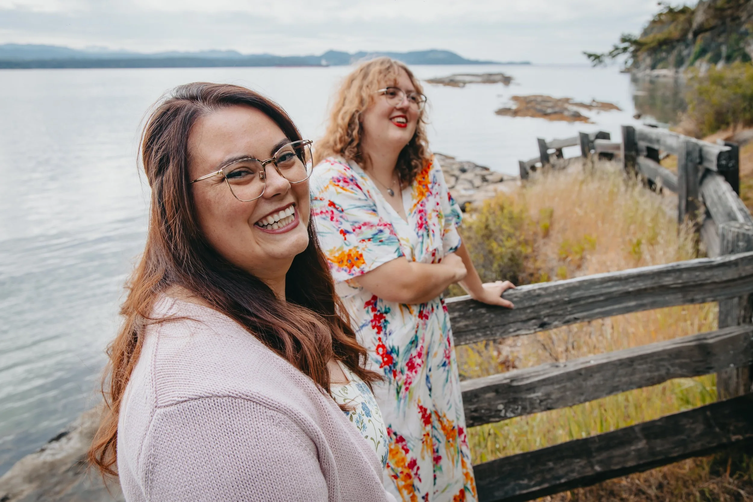 Two women smiling and standing near a wooden fence by the ocean, with trees and mountains in the background Outdoor portrait Photography on Gabriola Island BC