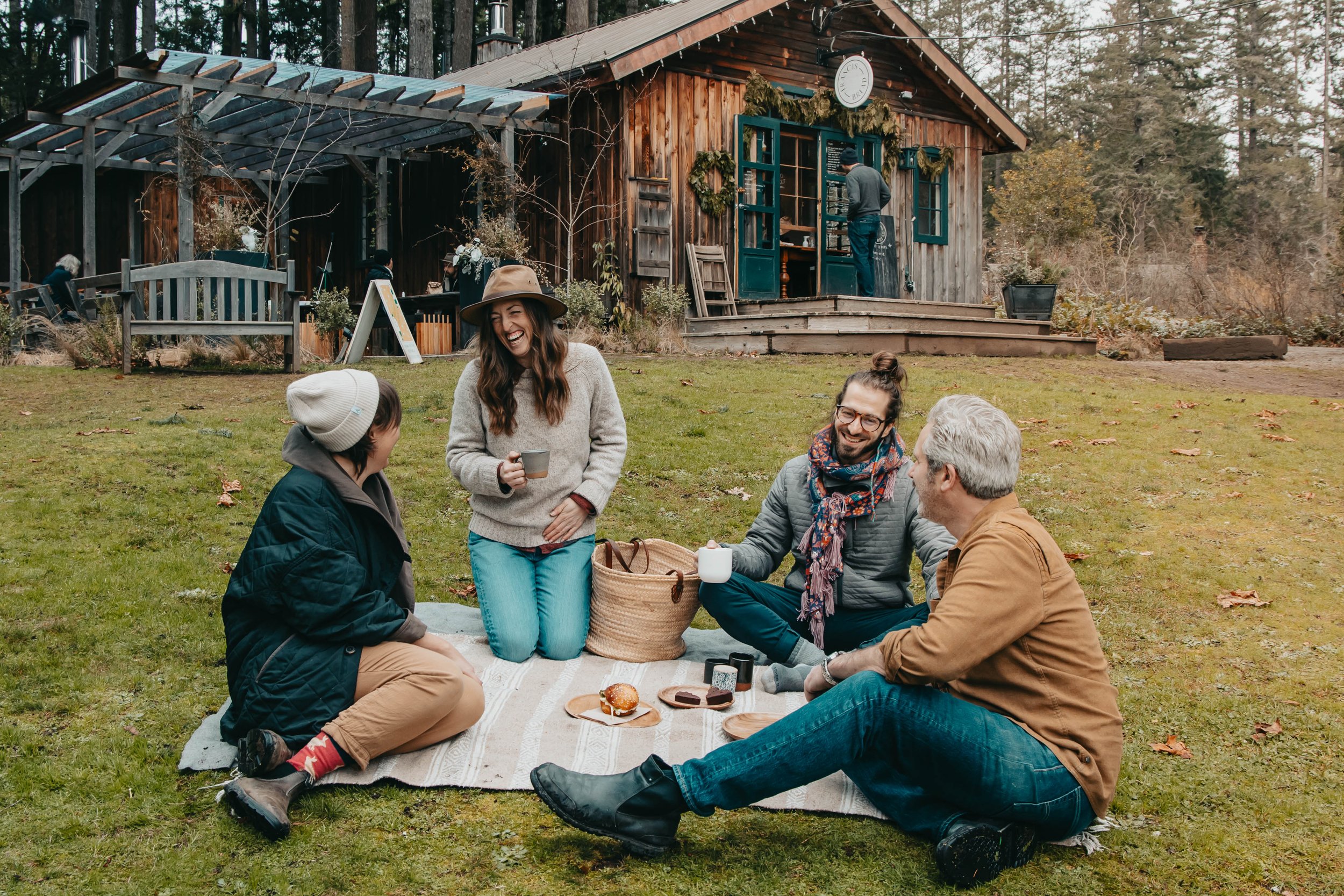 A group of friends enjoying a picnic outdoors on a blanket with food and drinks, in front of a rustic wooden house surrounded by trees.