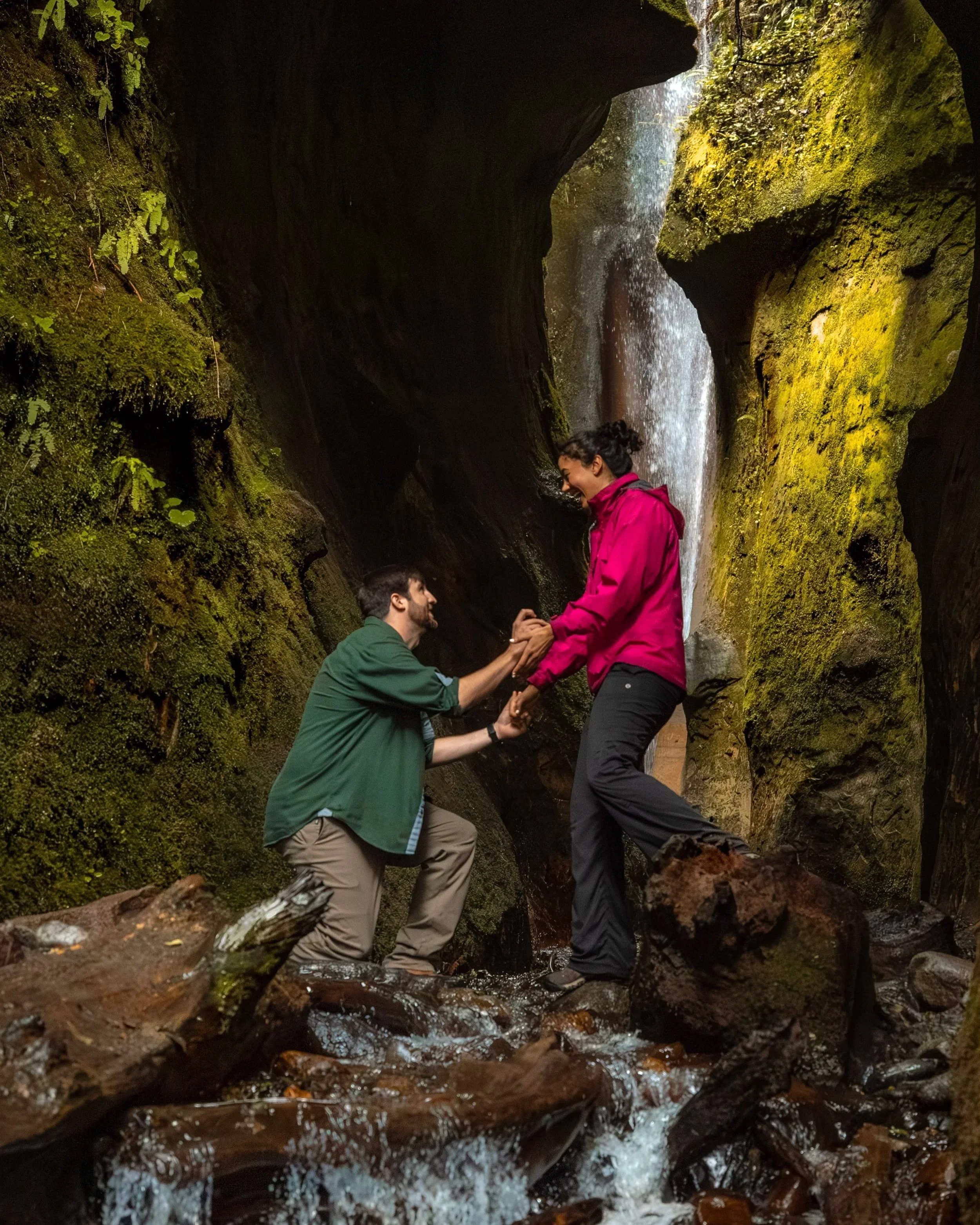 outdoor portrait of A man kneeling and proposing to a woman standing in a mossy canyon with a waterfall in the background, holding hands and smiling at a hidden waterfall at Sombrio Beach, Vancouver Island