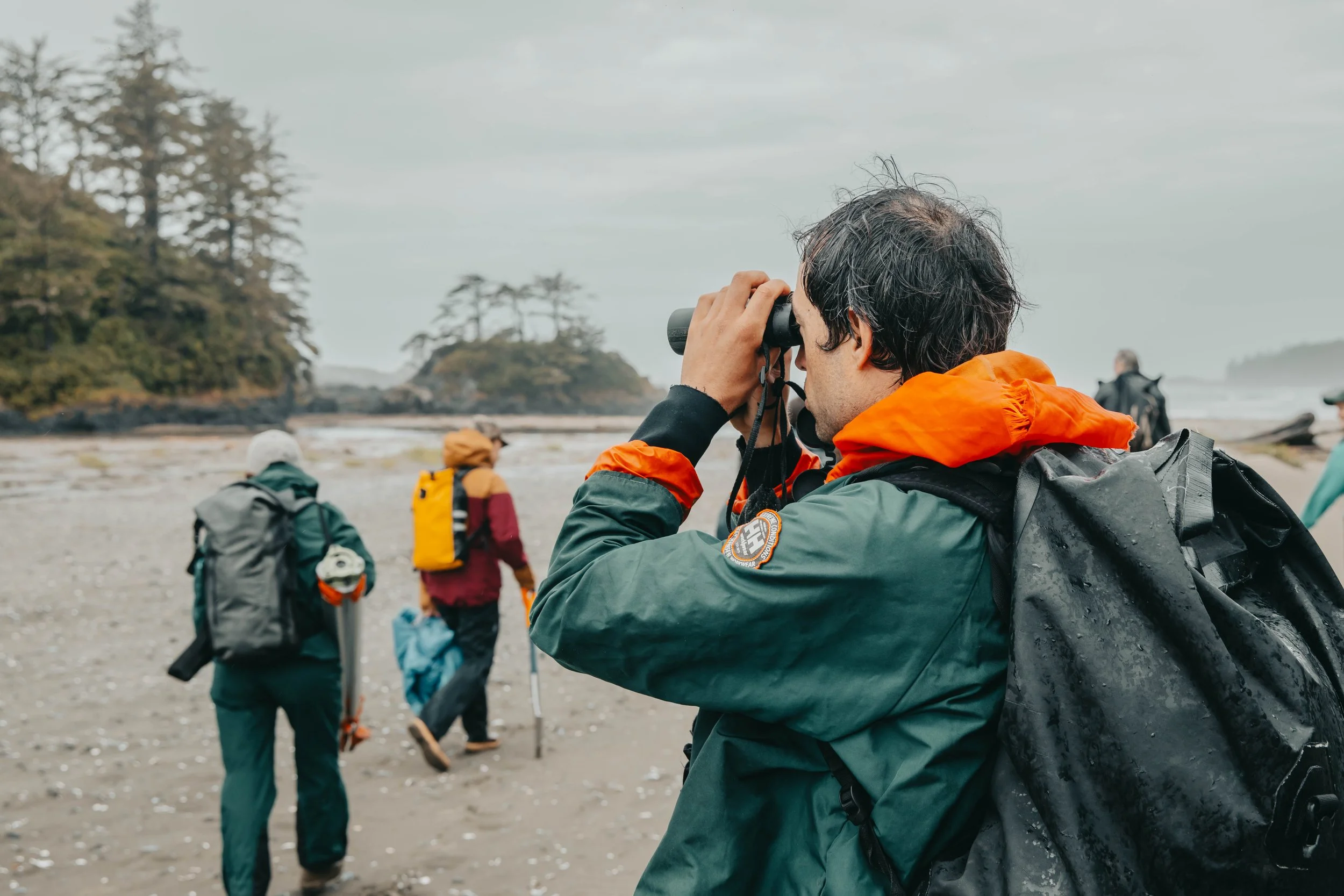 Group of hikers on a beach, some looking through binoculars, surrounded by trees and rocky terrain.