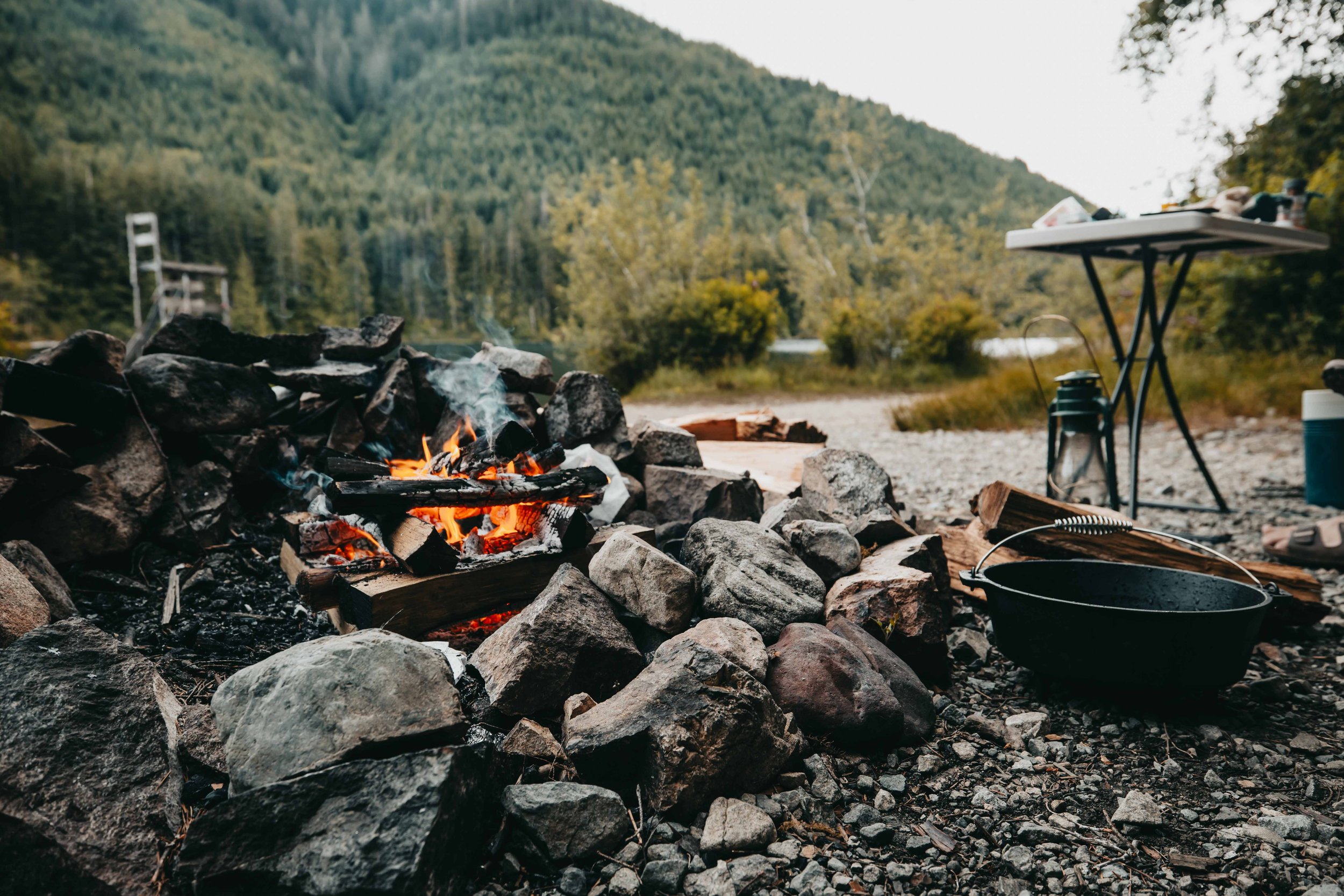 a campfire burns in a fire pit with a backcountry lake in the background. a cast iron pot and kerosene lantern sit next to the fire