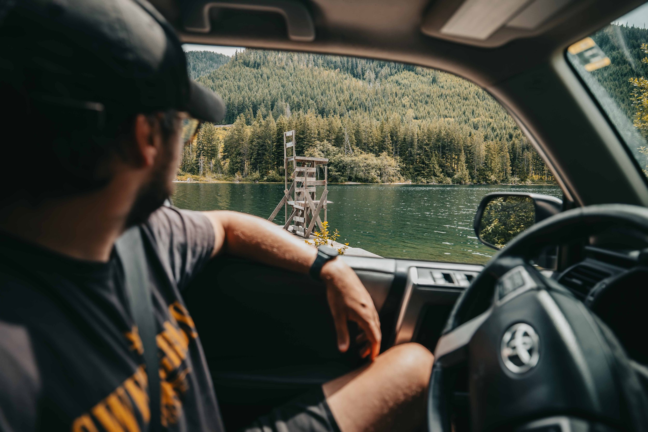 a man sits inside a toyota 4runner parked next to a lake gazing out the window at the green water and mountians