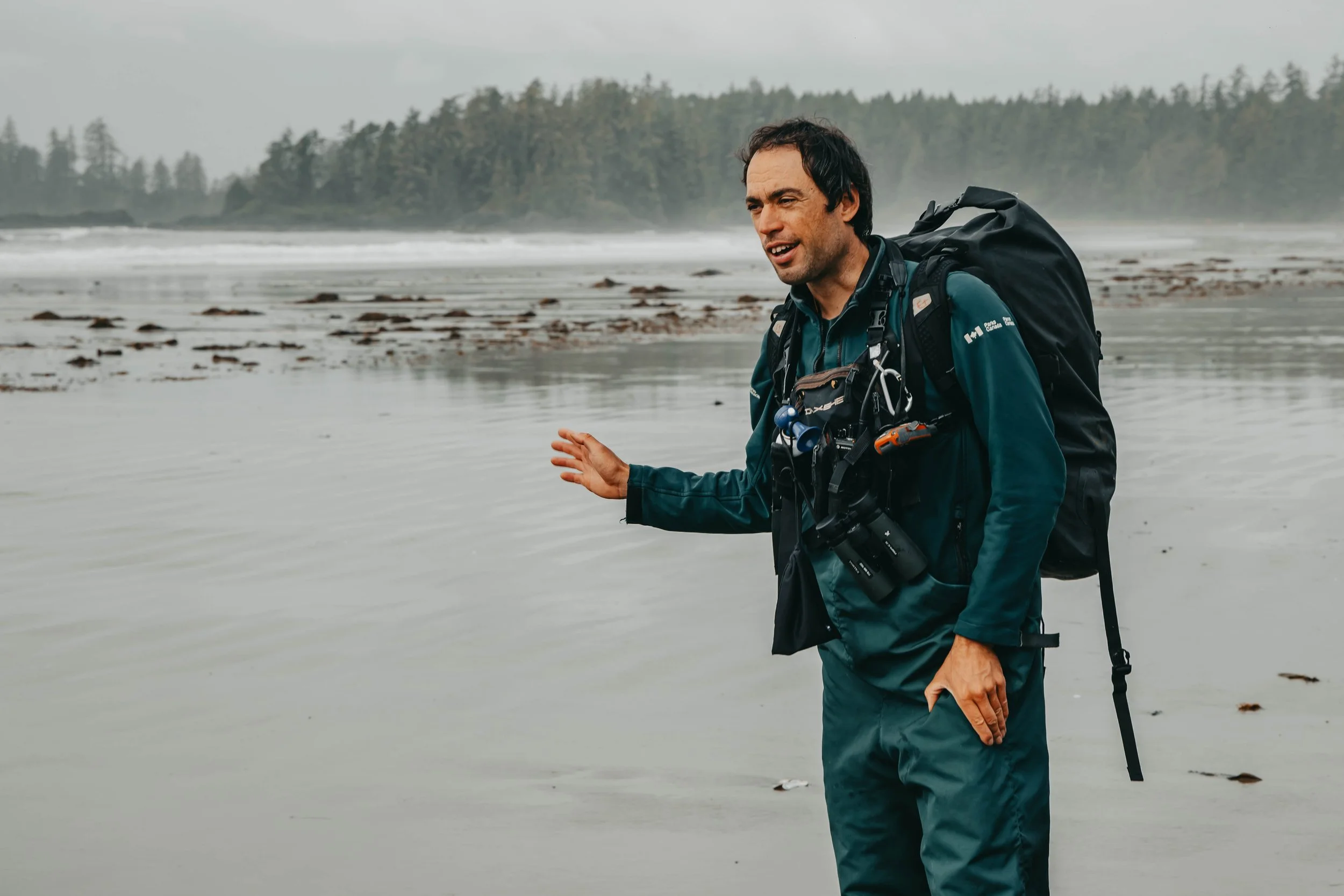 Man with a backpack on a beach, talking or explaining something, with a forested shoreline in the background on a cloudy day.