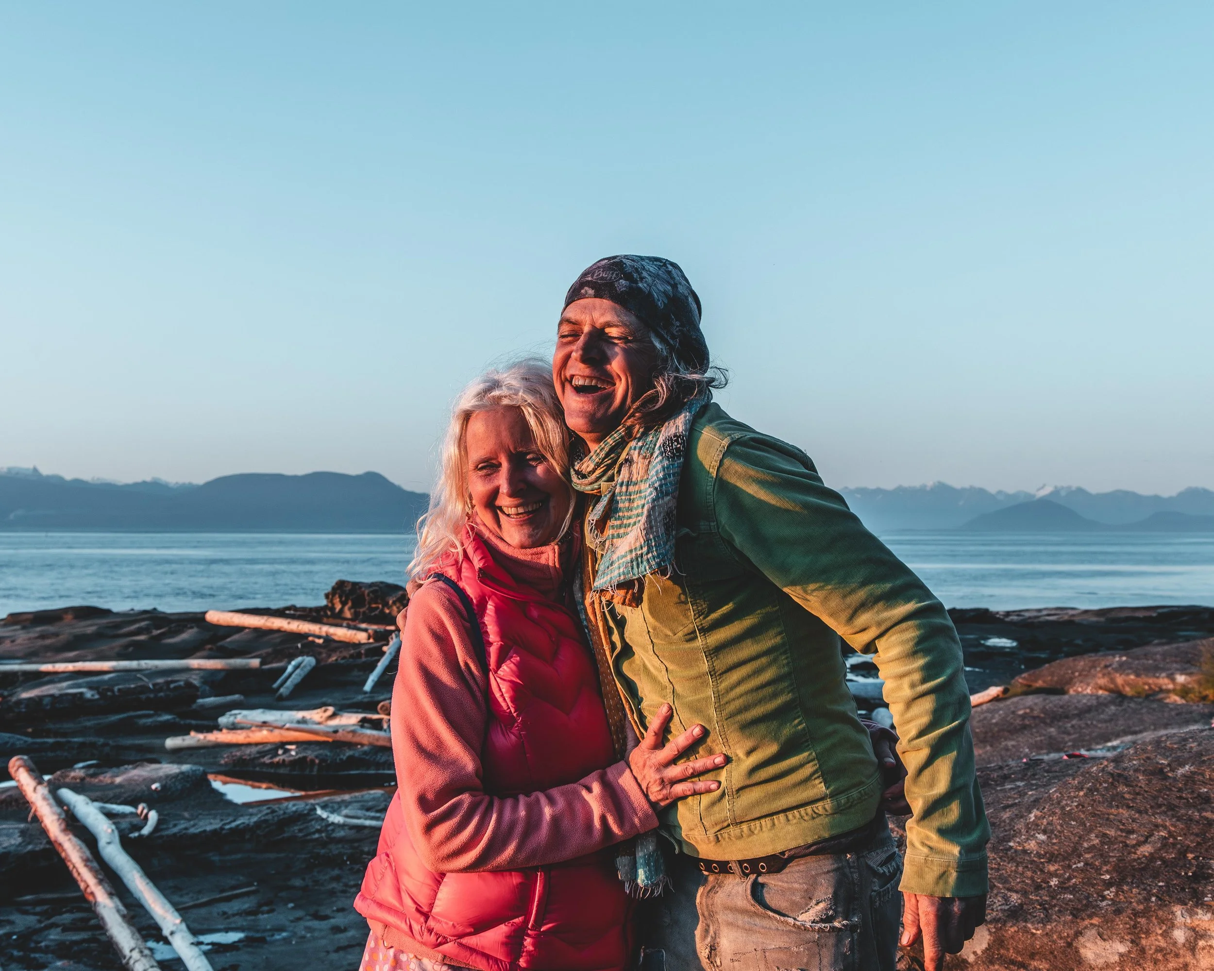 A joyful older woman and a man are hugging by the water with mountains in the background.