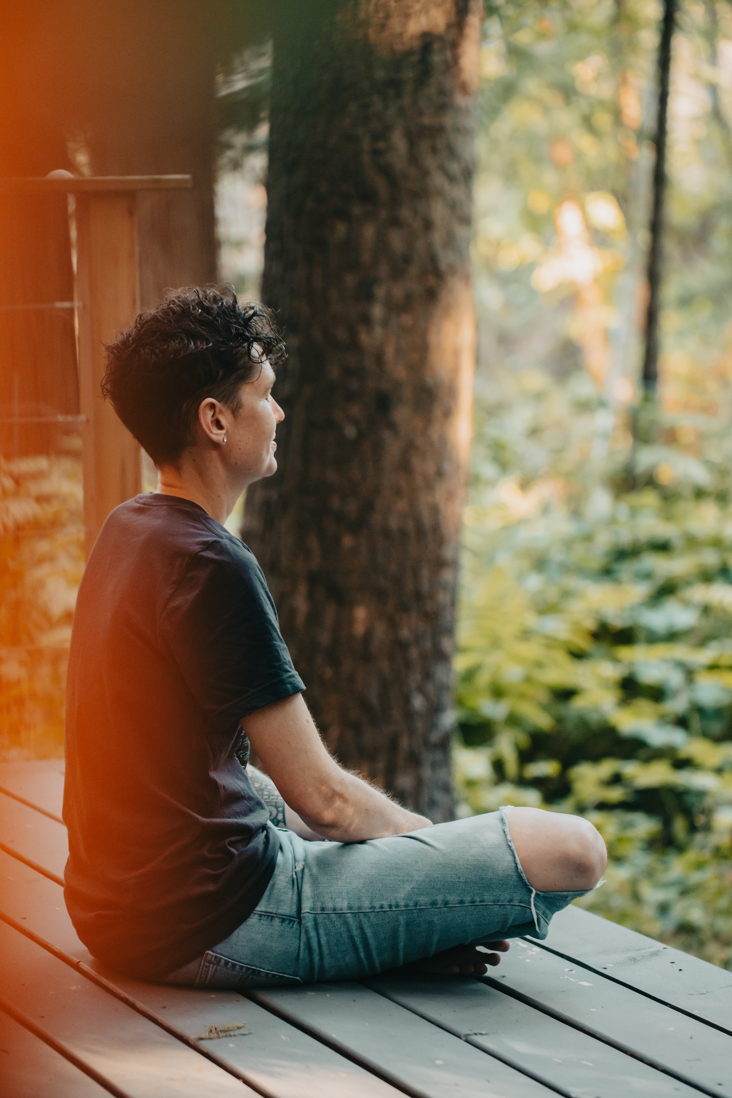 A woman with short curly dark hair in a black t-shirt and ripped jeans sitting cross-legged on a wooden deck in a forest, meditating or relaxing with her eyes closed Outdoor Portrait Photography on Gabriola Island BC