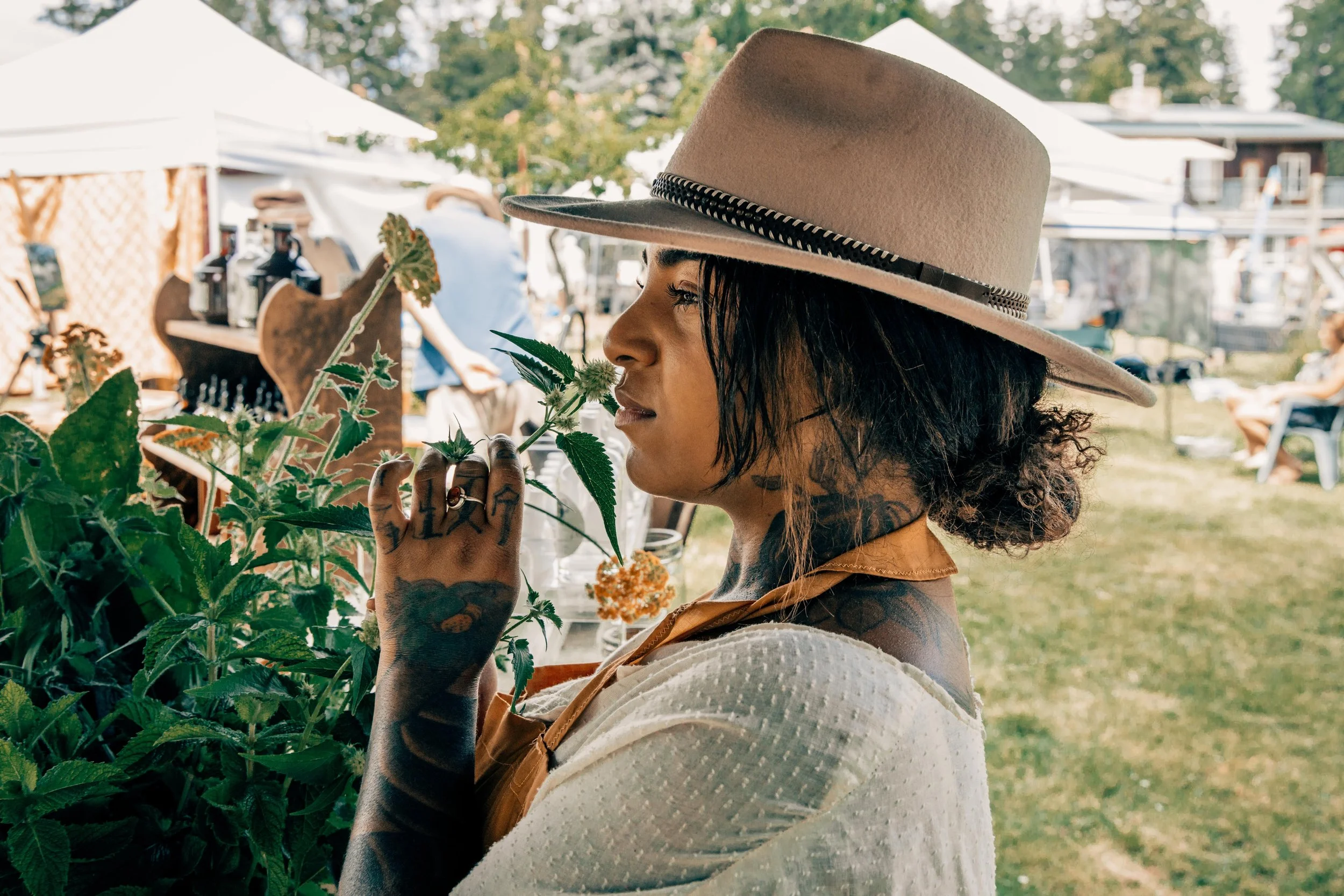 A woman with tattoos on her hand and neck, wearing a wide-brimmed felt hat with a decorative band, smelling a plant at an outdoor market or fair, with tents and people in the background.