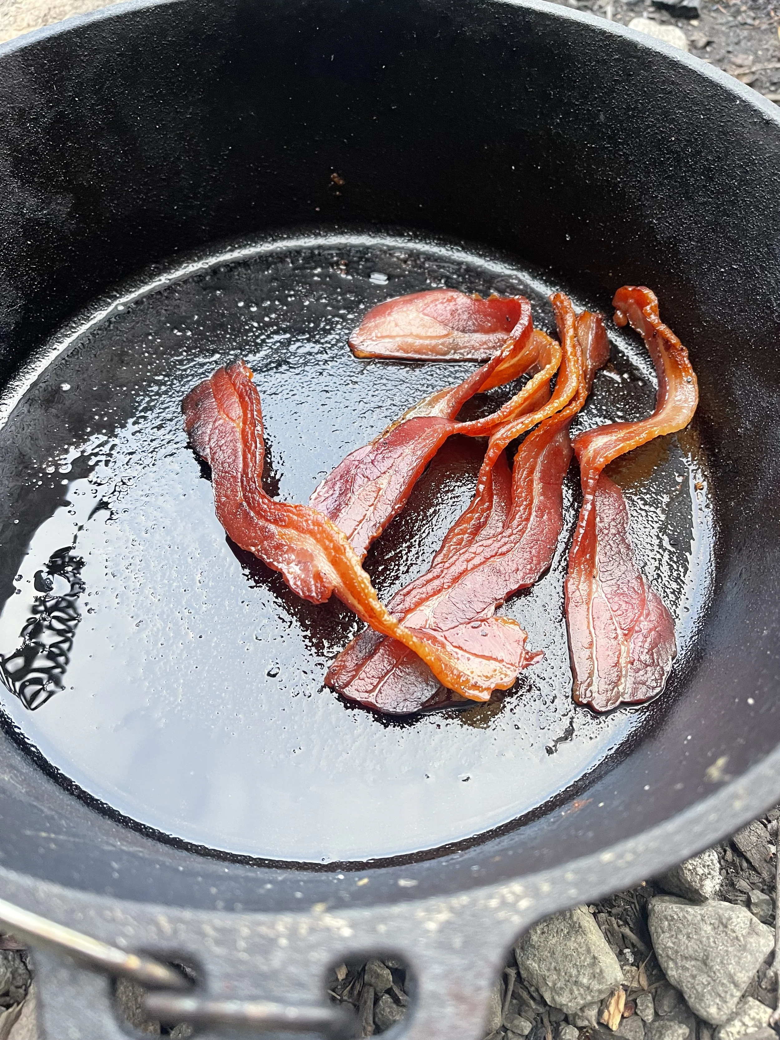 perfectly cooked bacon sits in a cast iron pot after being cooked on a campfire