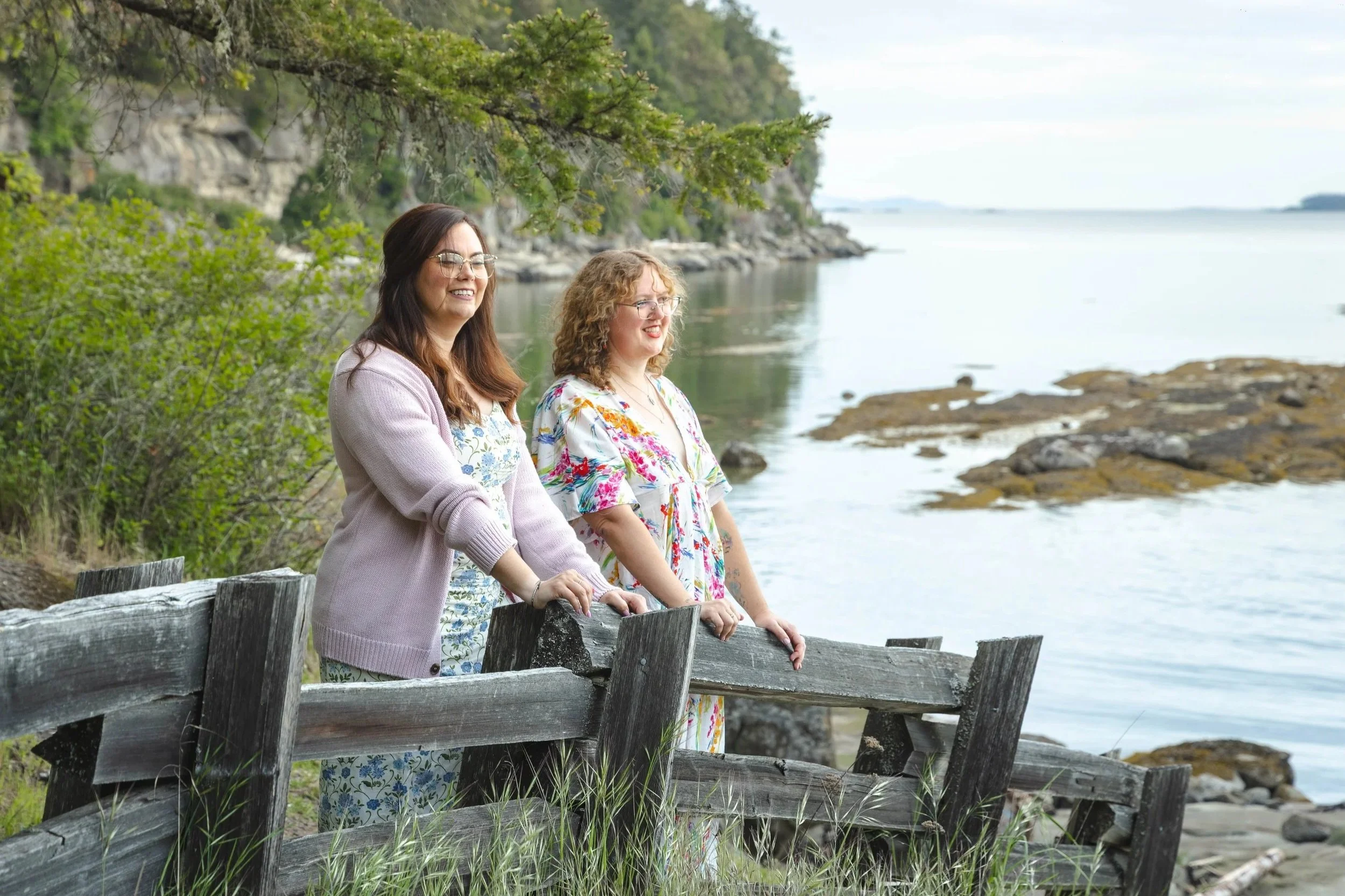 Two women standing behind a wooden fence near a rocky shoreline, smiling and looking out at the water with green trees and a cloudy sky in the background during outdoor portraits on Gabriola Island