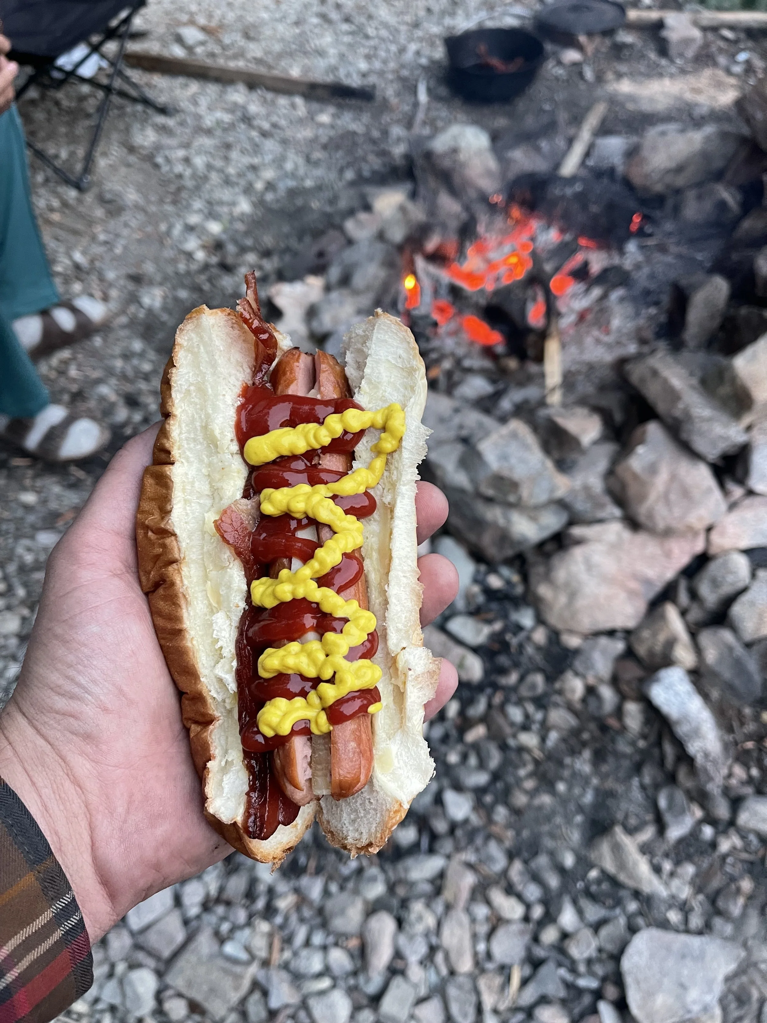 a hotdog covered in ketchup and mustard in the palm of a mans hand with a roaring campfire in the background