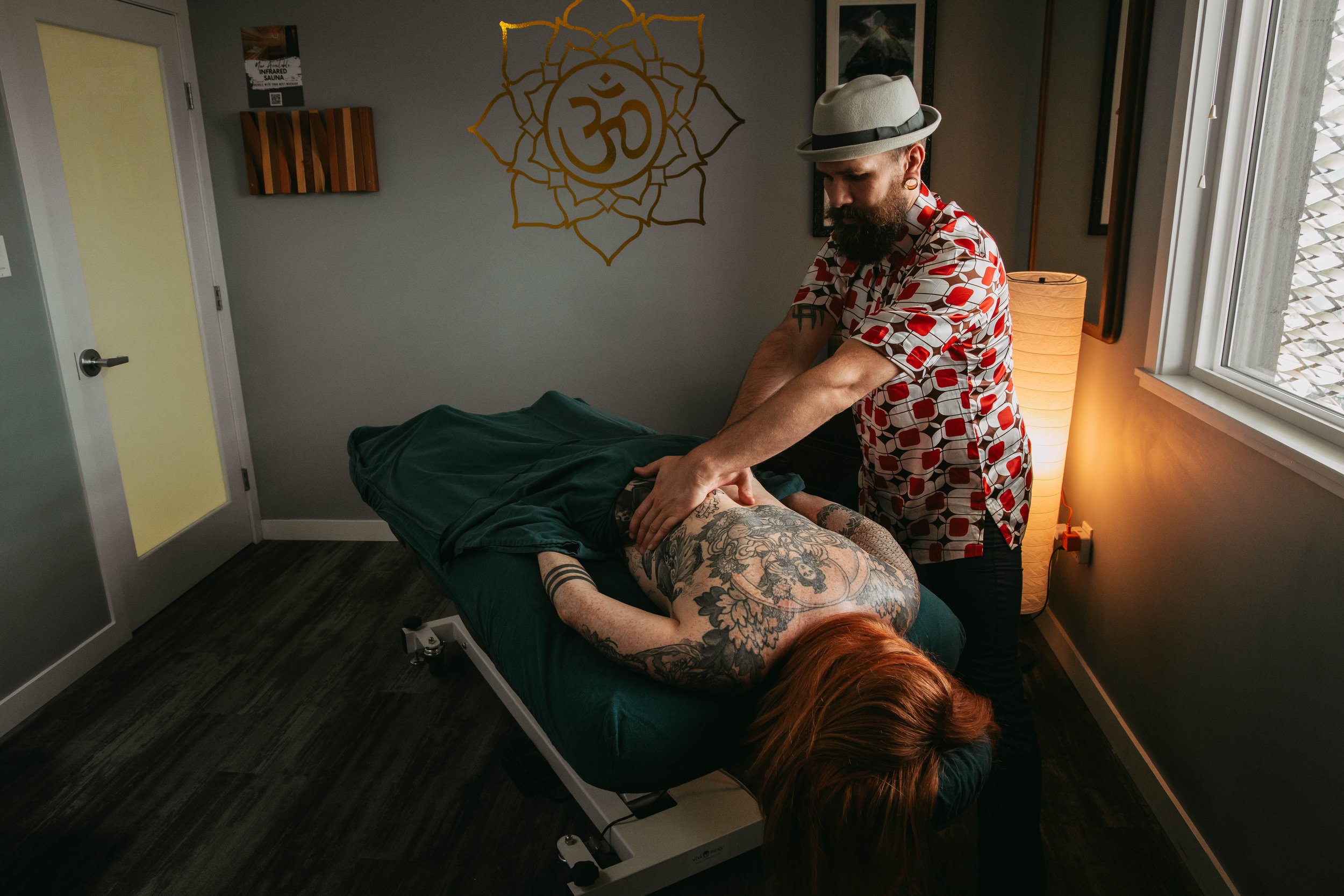 A person with red hair and tattoos receiving a massage from a man wearing a patterned shirt and a gray hat in a therapy room with decorative wall art and a window.