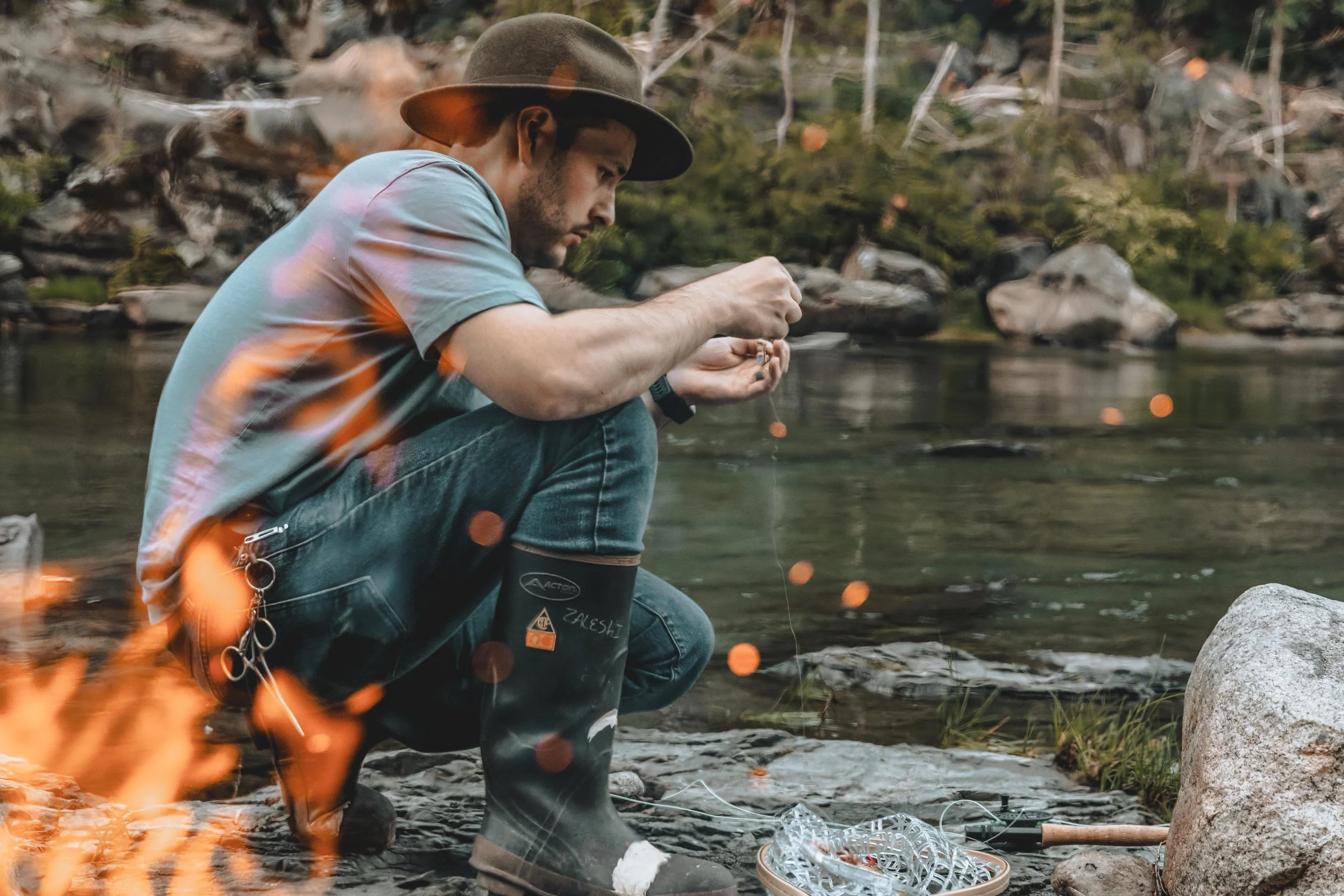 A man crouching by a river, wearing a hat, jeans, and rain boots, fishing with a fishing line. There is a fire burning in the foreground and a rocky landscape in the background.