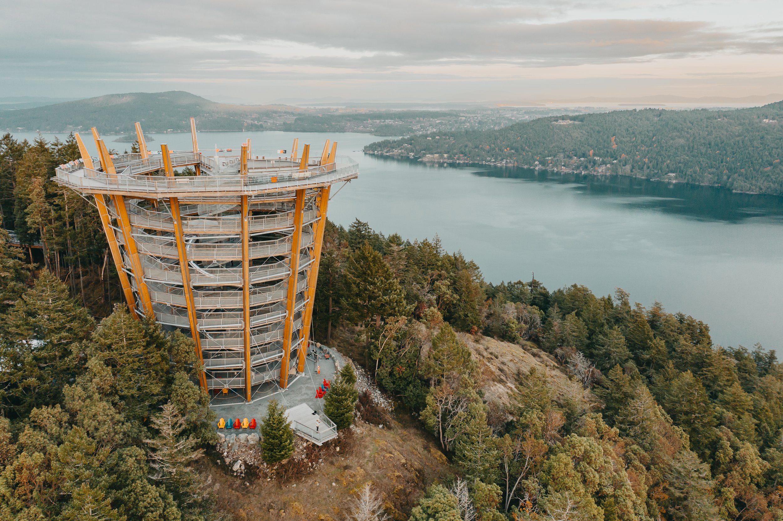 The Malahat skywalk, a tall, circular observation tower with a spiral slide on the outside, situated among dense trees on a hillside overlooking a lake and distant mountains under cloudy skies.