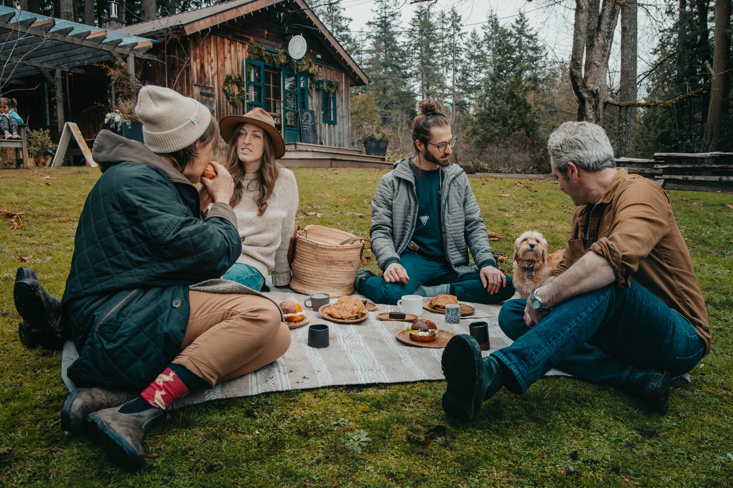 A group of four people and a small dog sitting on a blanket outdoors, having a picnic with food and drinks in front of a rustic wooden cabin forested background.