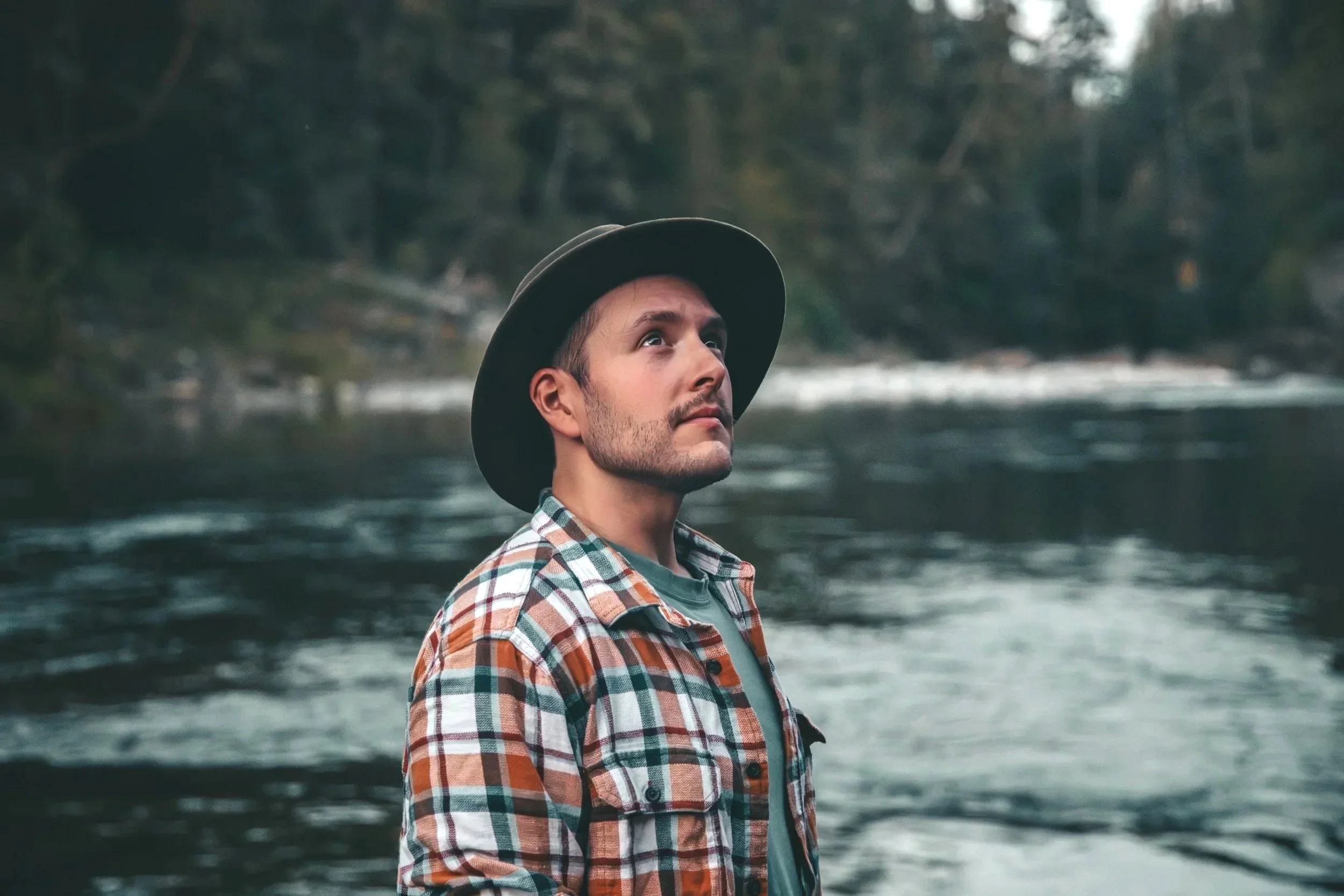 Outdoor Portrait of A man wearing a plaid shirt and a wide-brimmed hat standing near a river or lake surrounded by trees