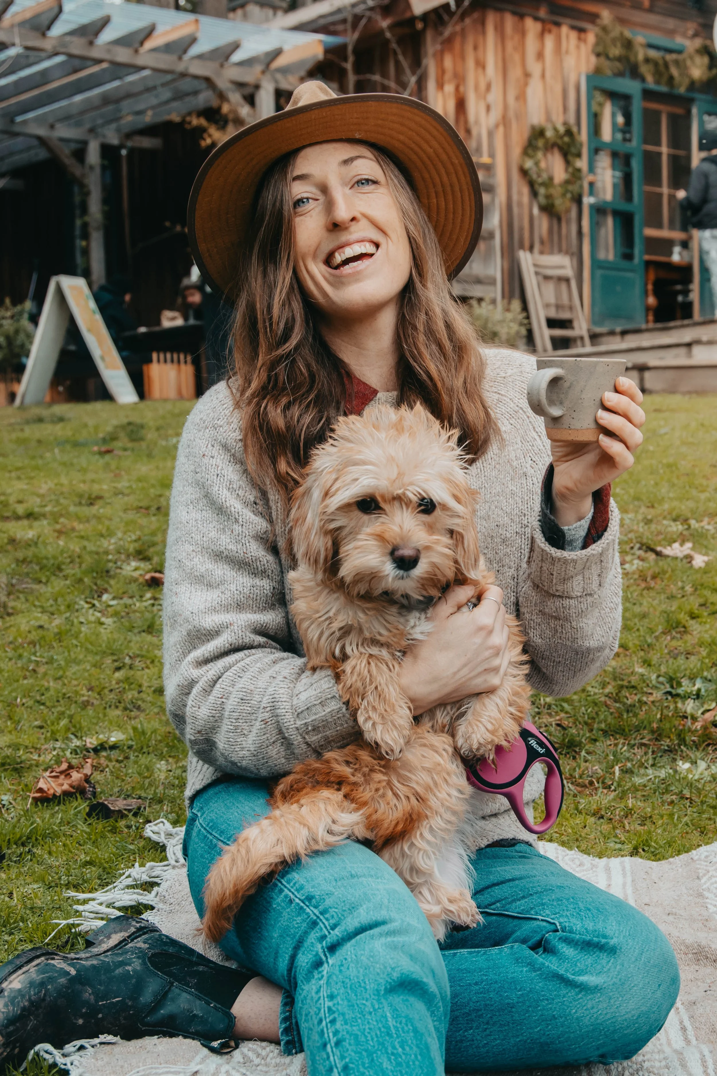 A woman wearing a wide-brimmed hat and a cozy sweater sitting on a blanket outdoors, holding a fluffy dog and a ceramic mug, with a rustic wooden building in the background.