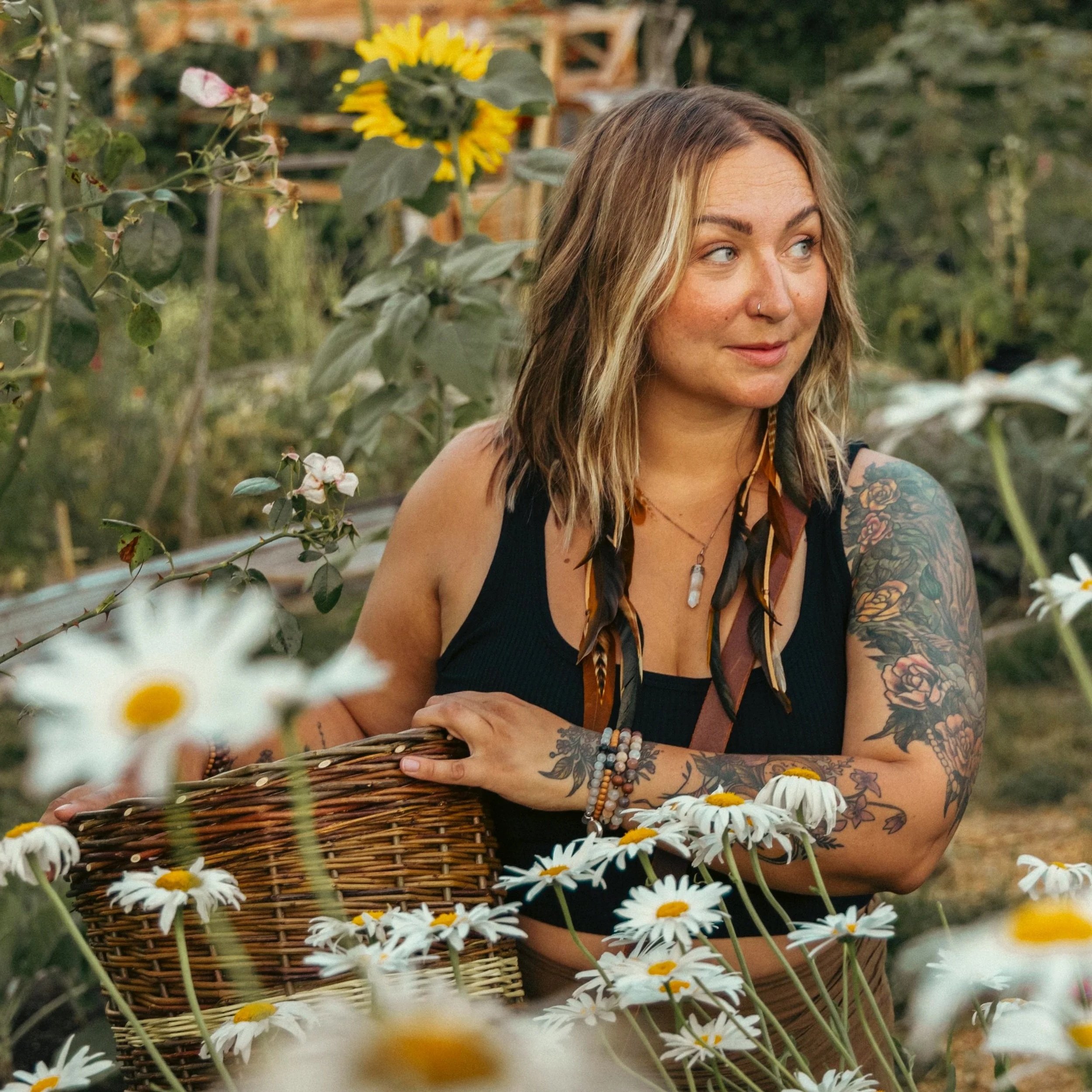 A young woman with tattoos on her arm, wearing a black tank top and beaded bracelets, is in a garden gathering daisies with a wicker basket. She has wavy, light brown hair with blonde highlights and is smiling slightly while looking to her left. There are yellow sunflowers and greenery in the background.