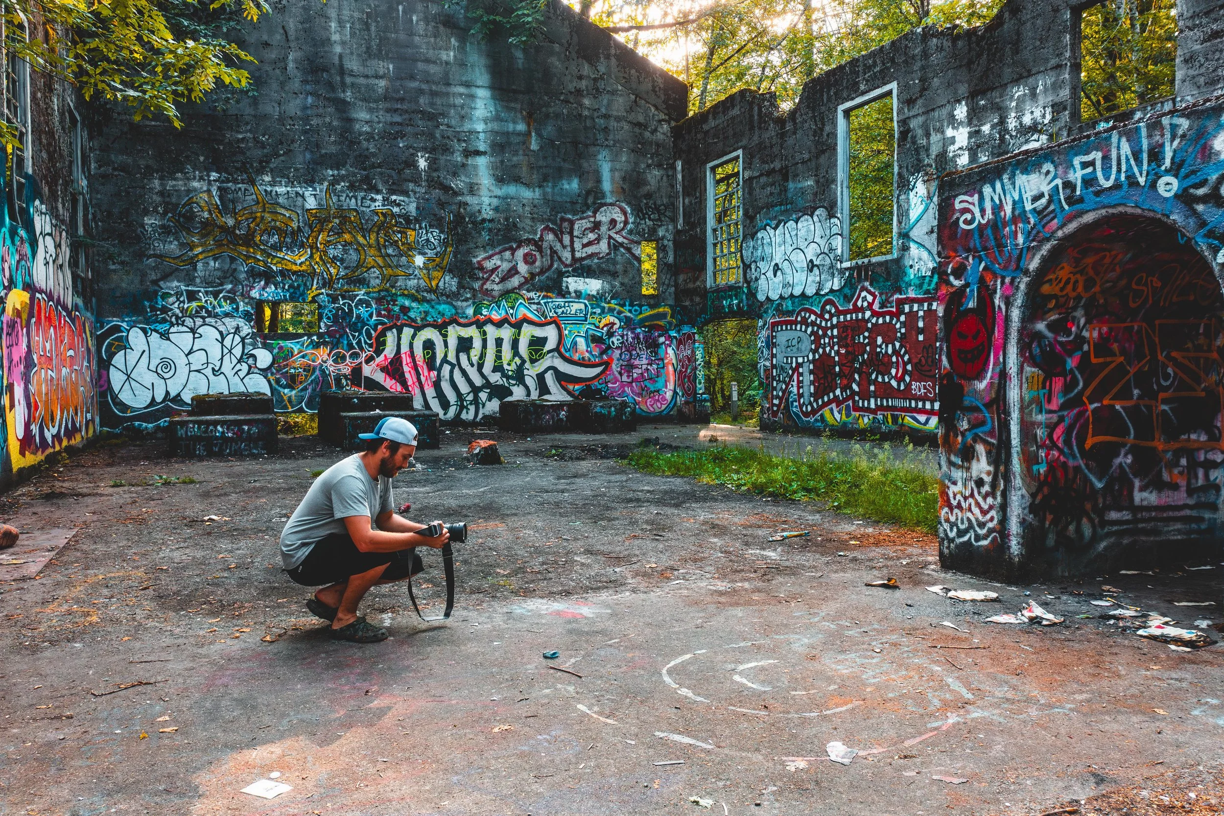 A person in a grey t-shirt, black shorts, and a backwards cap crouches on the ground taking a photograph with a camera inside an abandoned, graffiti-covered building. The building is surrounded by greenery, and sunlight shines through the window openings.