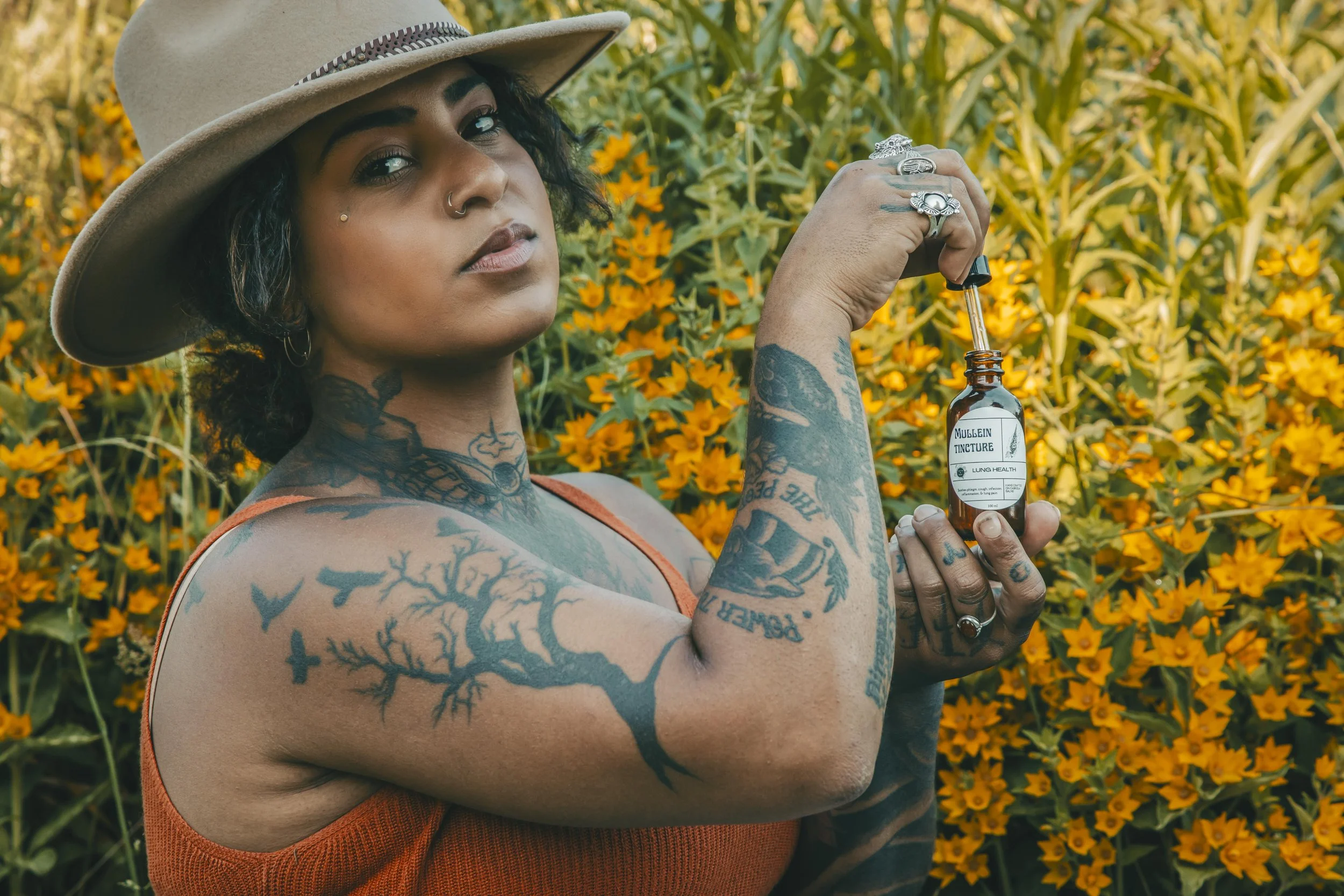 A tattooed woman wearing a beige hat with a silver band, holding a small brown bottle labeled 'Mullein Tincture' with a dropper, standing outdoors amidst yellow-orange flowers.