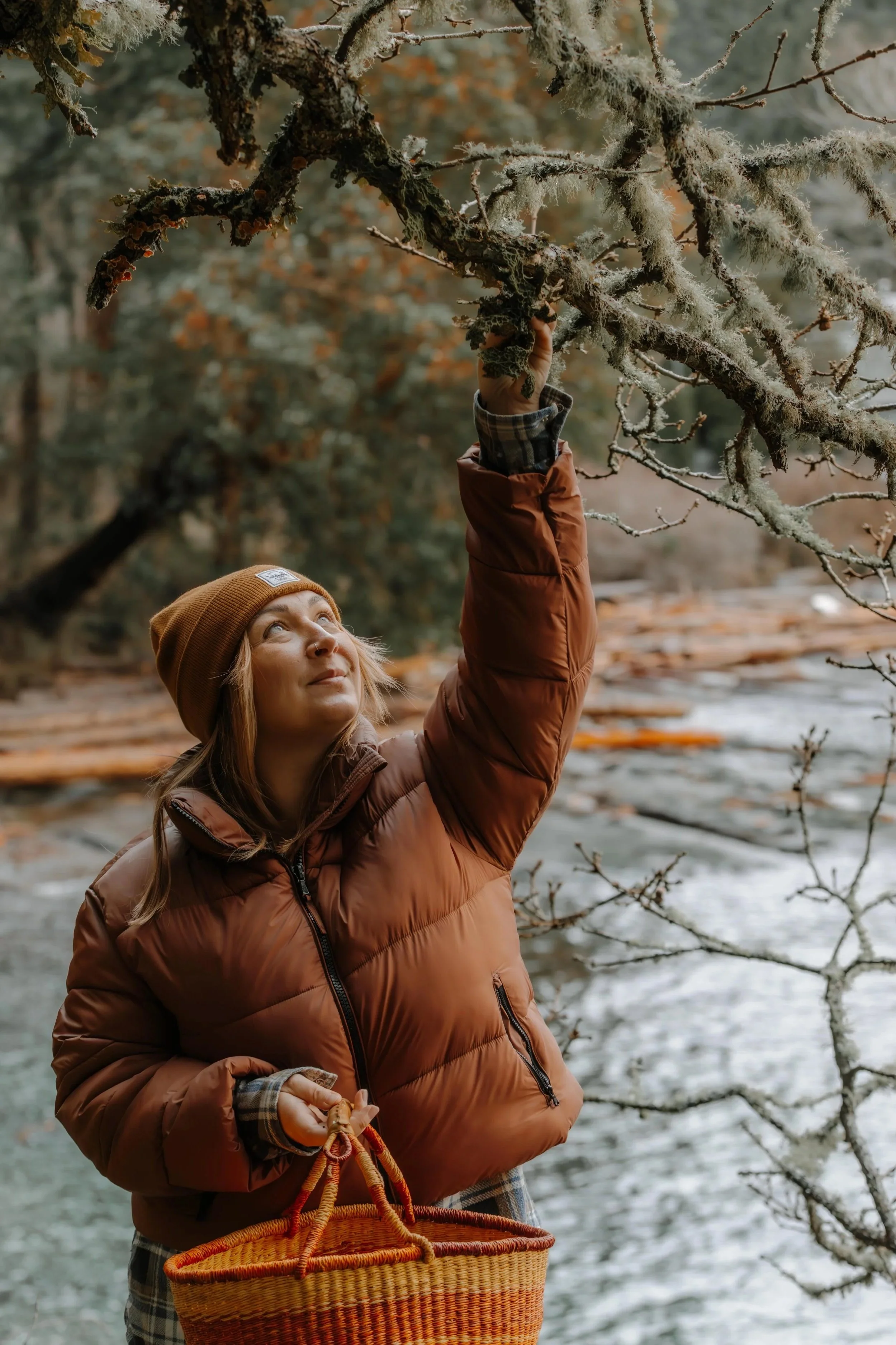 A woman in a brown jacket and hat holds onto a branch covered in moss and lichen in a forest near a river, carrying a wicker basket.