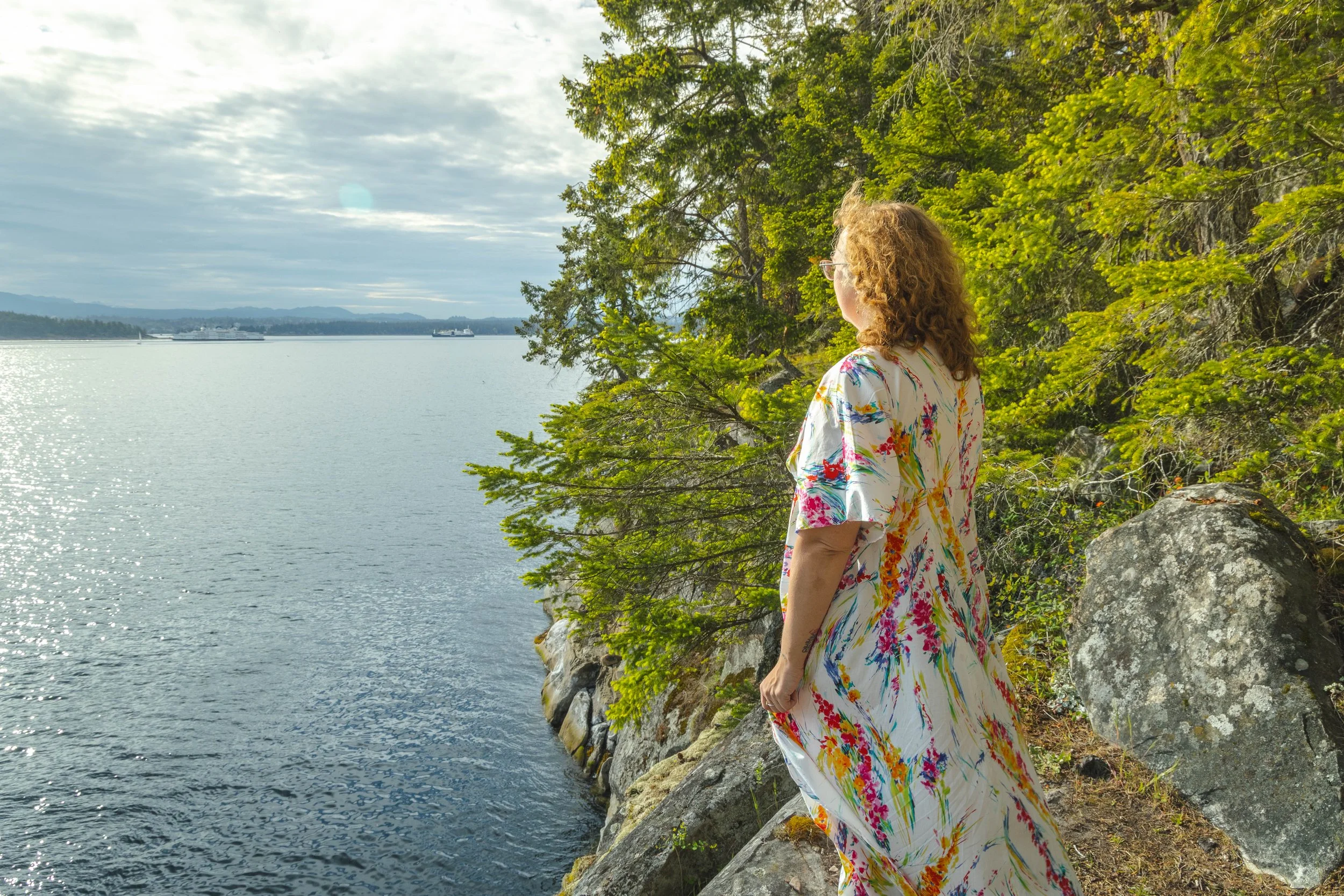 Outdoor portrait of A woman with curly red hair wearing a colorful floral dress standing on a rocky shoreline looking out over a body of water, with trees and distant ships visible on Gabriola Island BC
