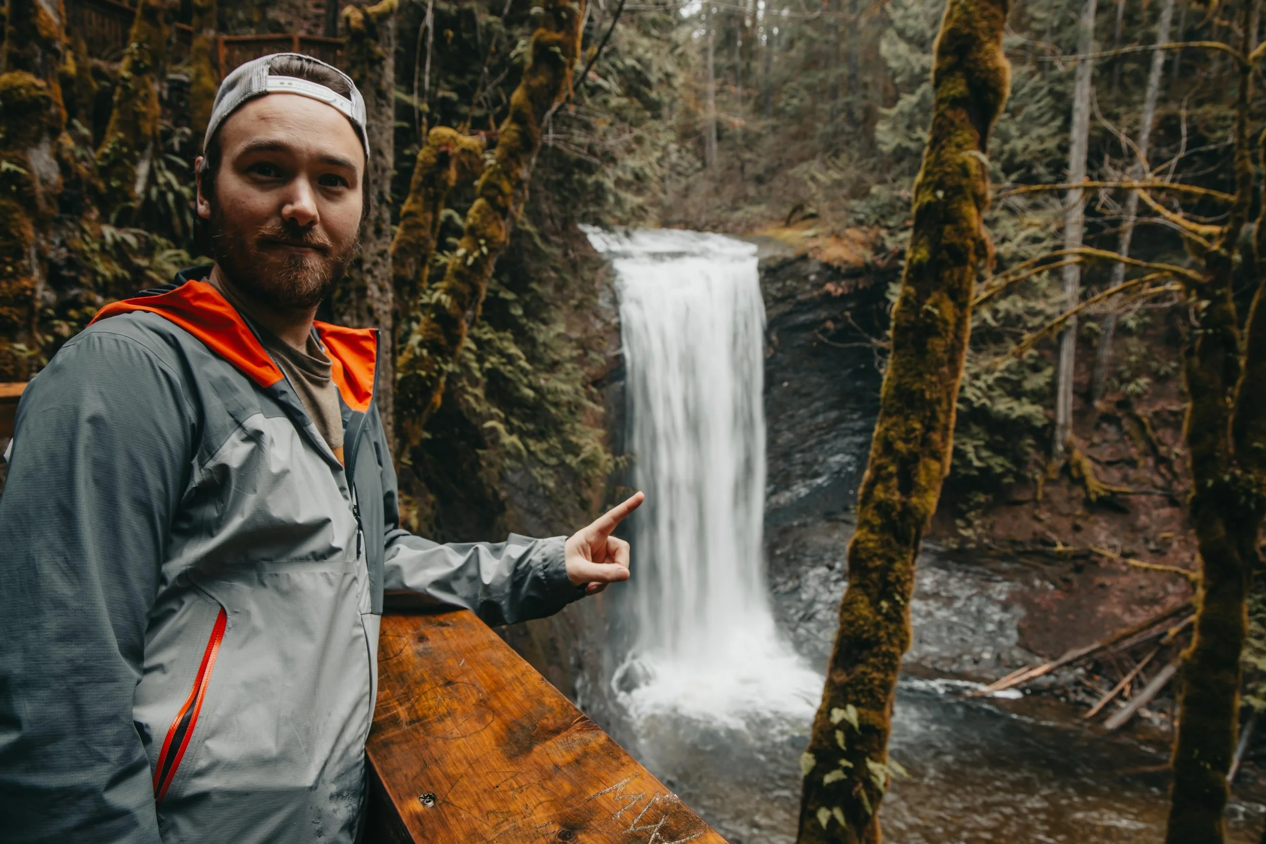 A man with a grey jacket and a backwards cap pointing at a waterfall in a forested area ammonite falls, nanaimo BC