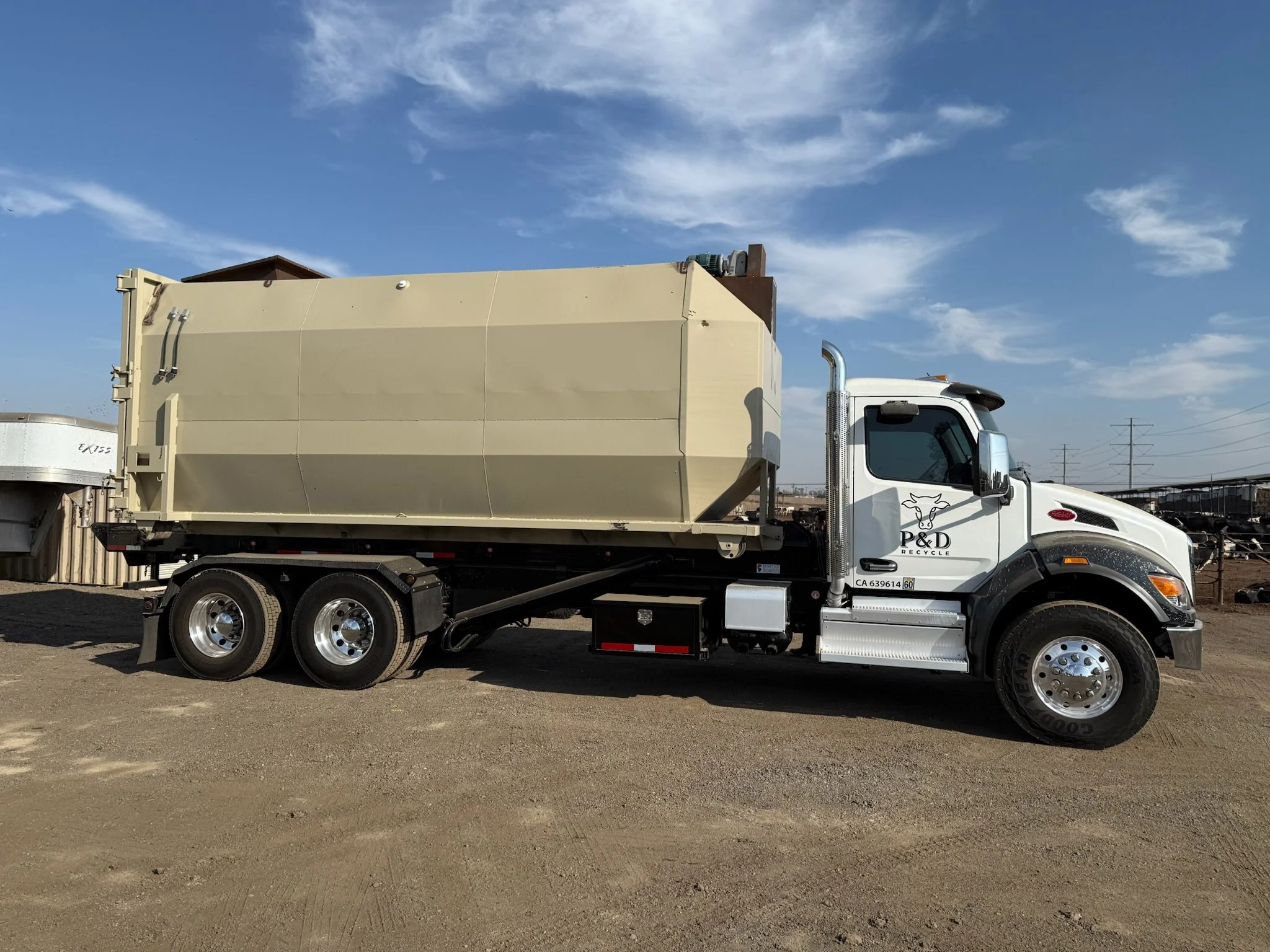 A white semi-truck with a beige cargo container parked on a dirt lot under a blue sky with some clouds.