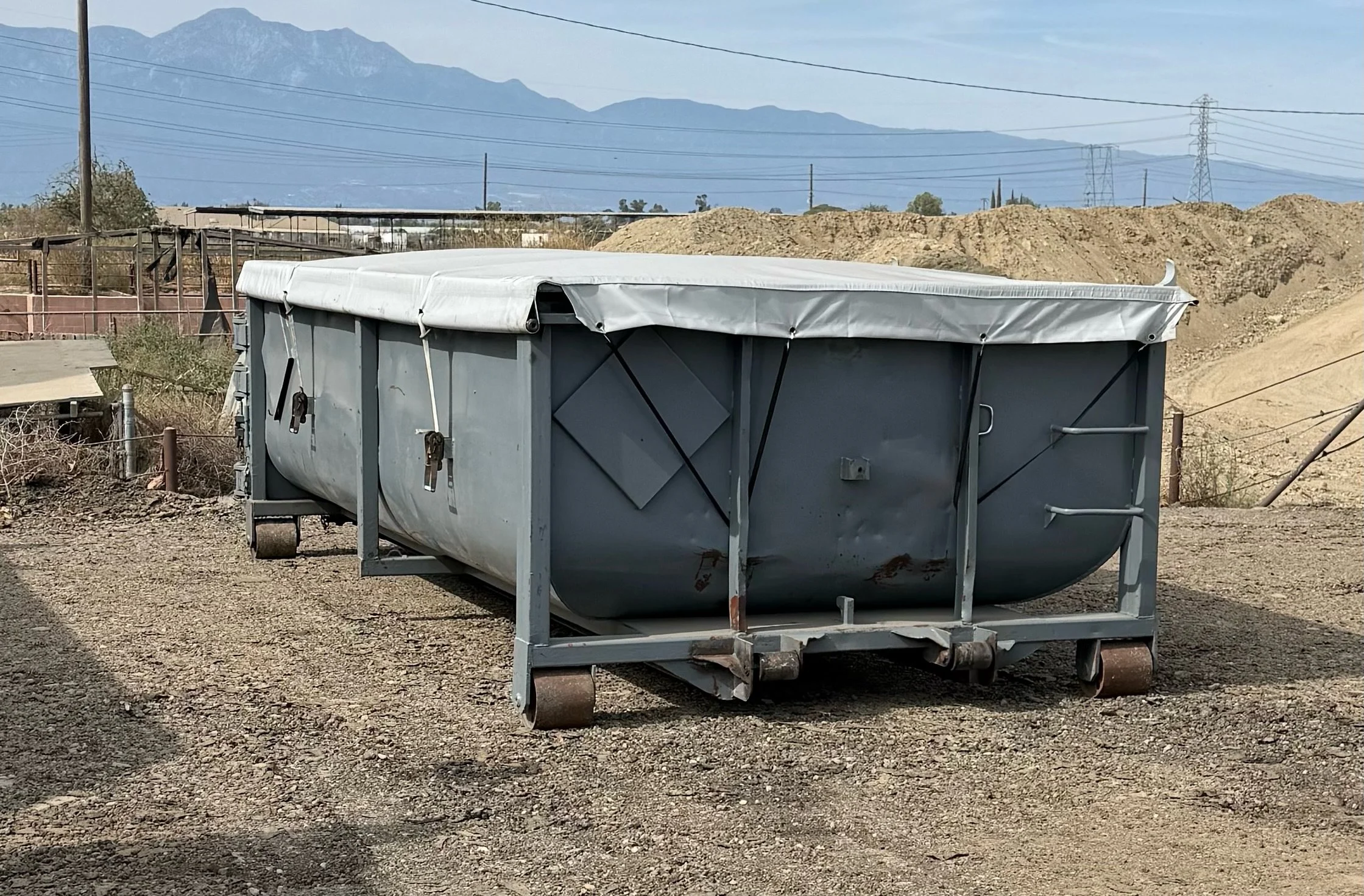 A metal dumpster with a white cover in a dirt lot, with power lines, mountains, and clear sky in the background.