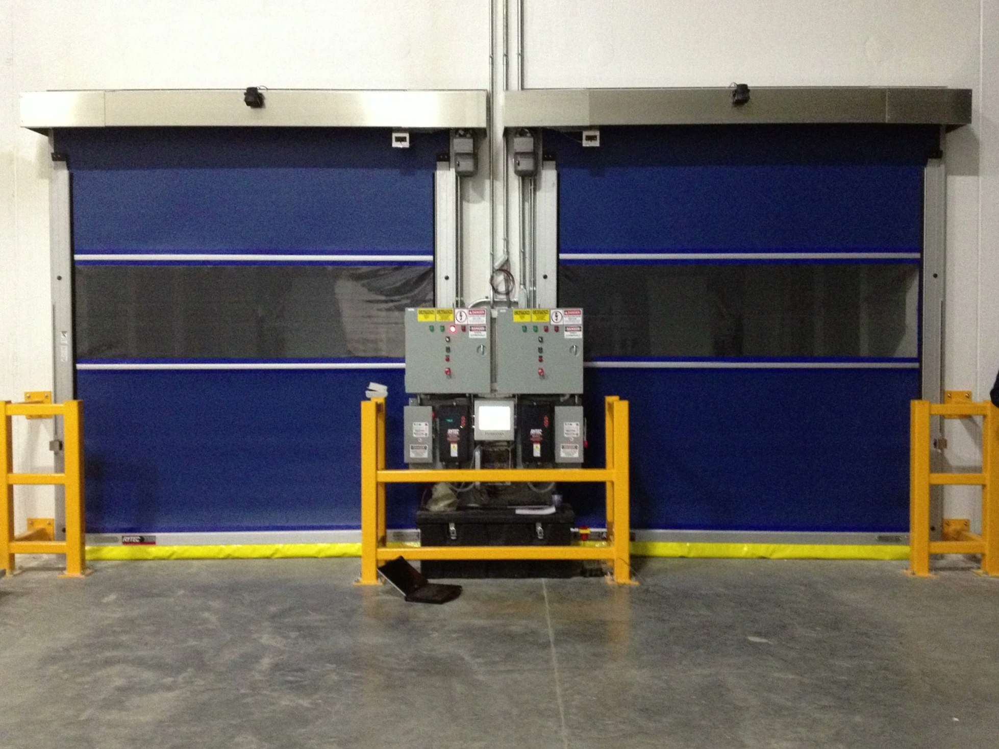 Industrial setting with two large blue and gray machinery doors, yellow safety barriers, control panel with indicator lights, and electrical wiring.