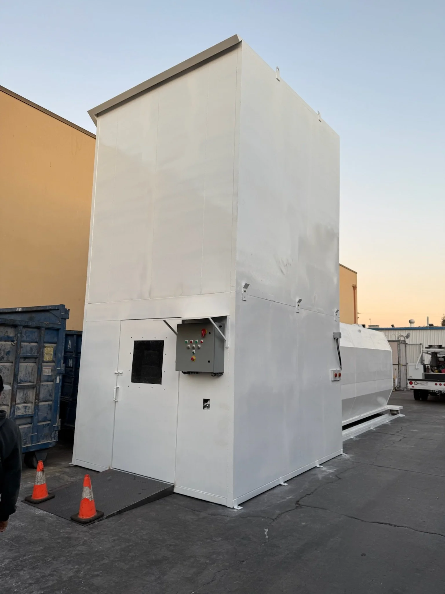 Large white industrial machine with control panel, situated outdoors on pavement, with orange traffic cones nearby and a beige building in the background at dusk.