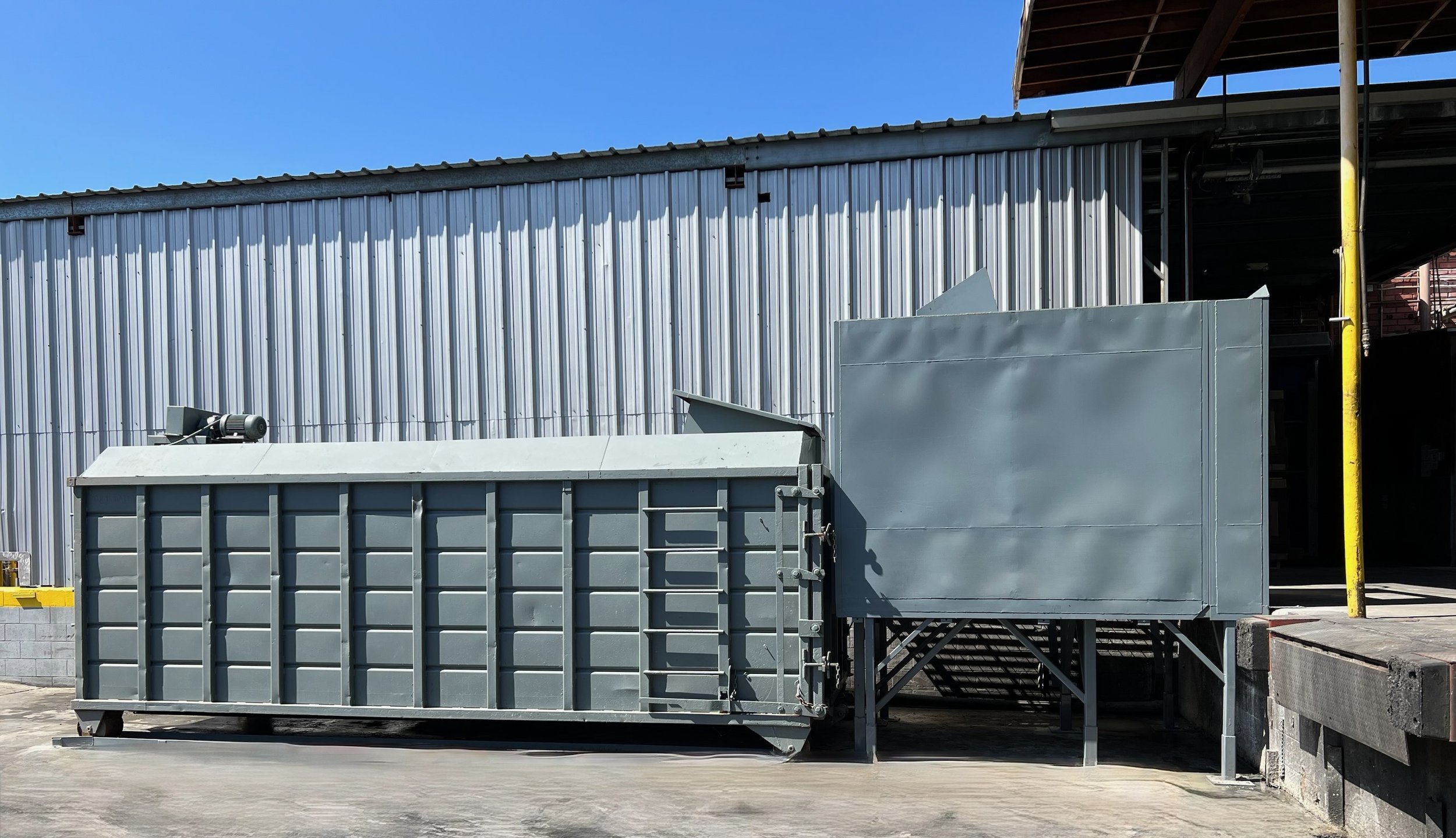 Industrial scene with metal equipment, a large grey storage container, and a corrugated metal building under a blue sky.