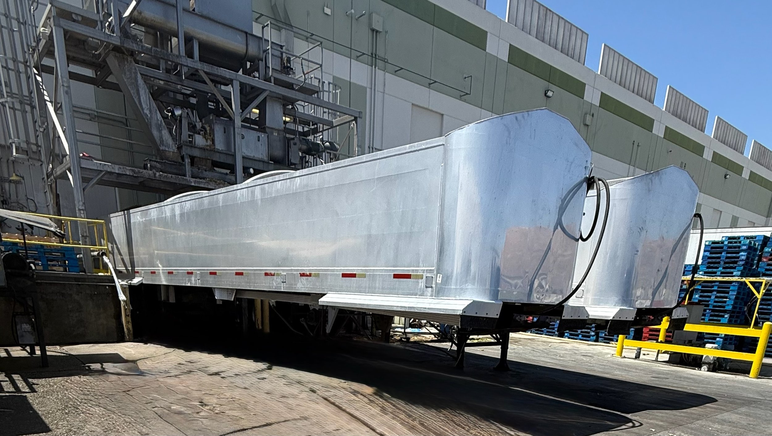 A large industrial metal trailer parked outside a building, with scaffolding and industrial equipment visible in the background, and blue sky overhead.
