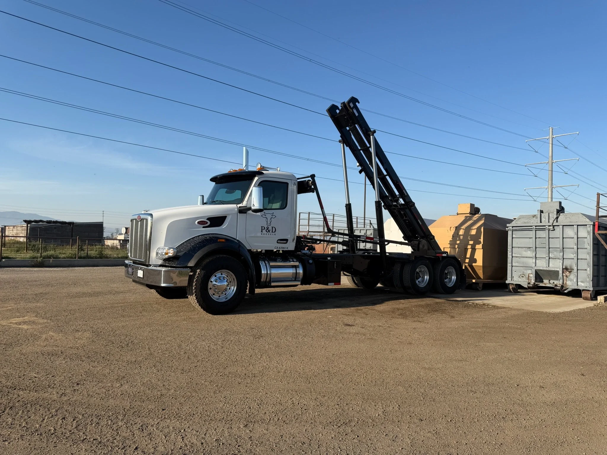 A white truck with a crane attachment on it, parked on a dirt lot with some buildings and power lines in the background.