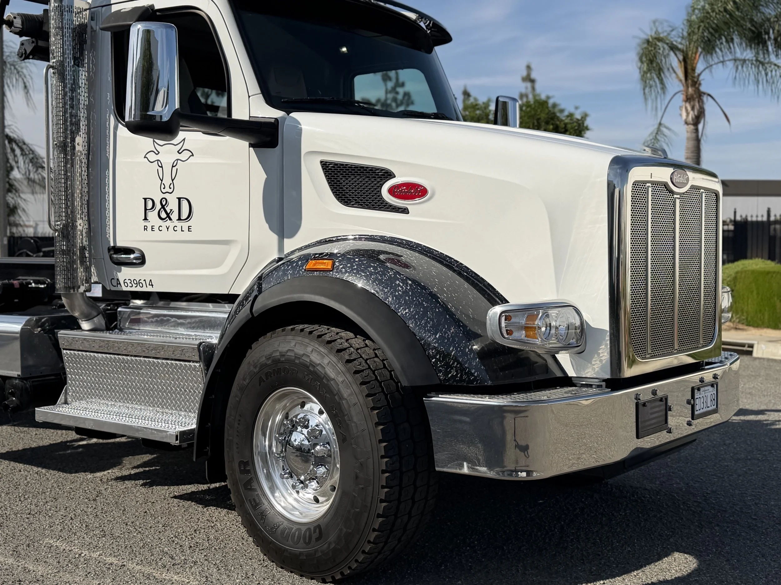 A white Peterbilt truck with a logo of a cow and the words P&D Recycle on the door, parked outdoors with palm trees and a fence in the background.