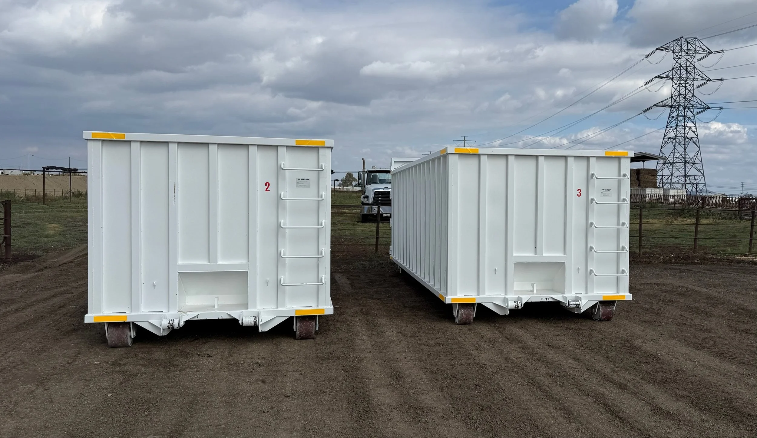 Two white industrial equipment containers with the number 2 and 3 written on them, placed outdoors on brown dirt ground, with a truck in the background and large electrical power line towers under a cloudy sky.